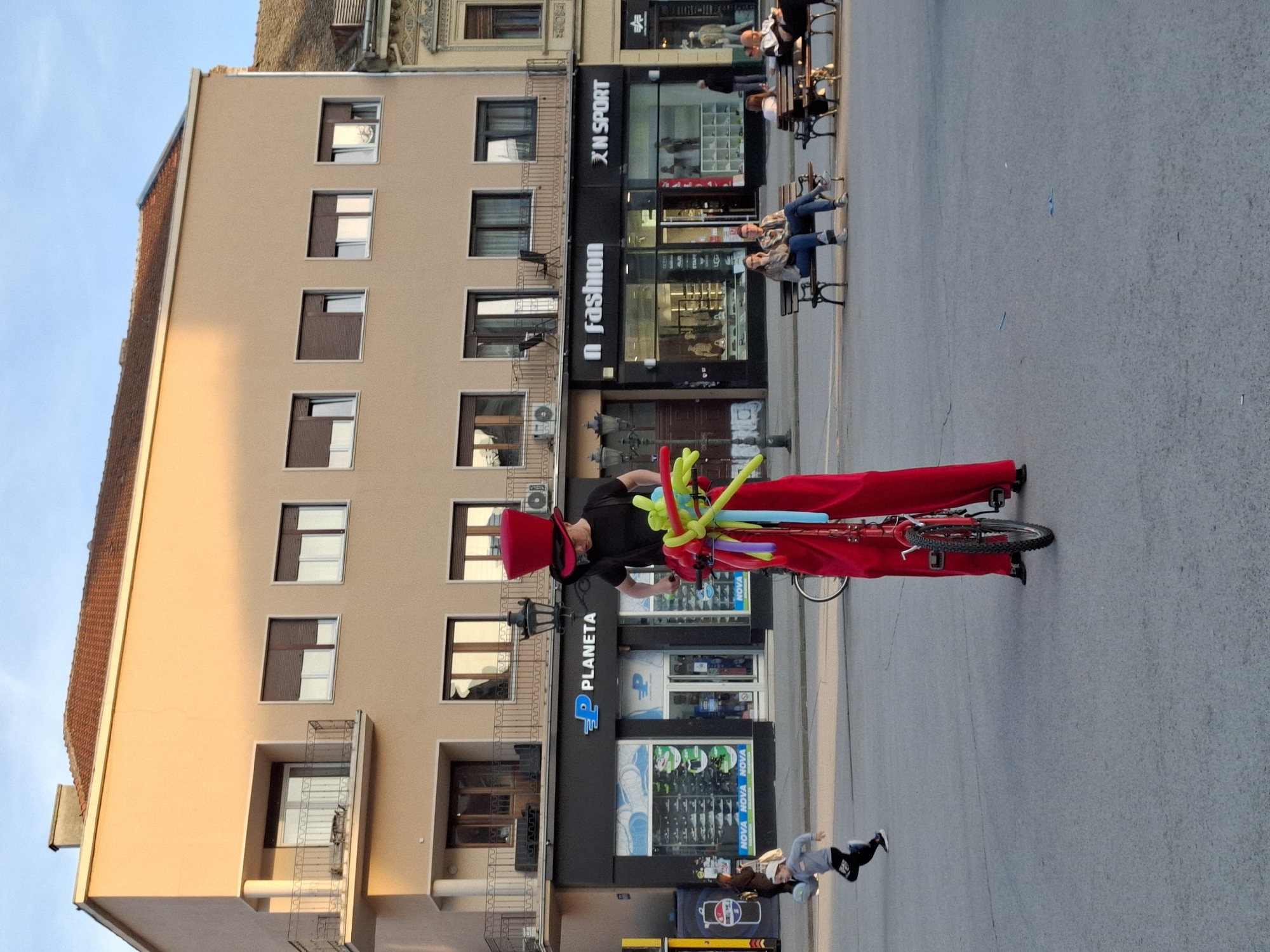 A man wearing a large red hat and black shirt is riding a red bicycle on a city street in Novi Sad, Serbia. The bicycle is decorated with colorful balloons. He is in front of a building with several businesses, including "Planeta," "N fashion," and "X N Sport." People are sitting on benches in front of the stores. A pedestrian is walking on the sidewalk. The building is multi-story with multiple windows and some small balconies. The street is wide and paved.