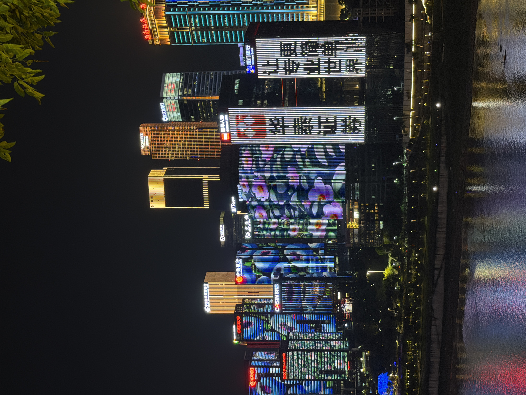 This image depicts a nighttime cityscape in the Yuzhong District of China. A body of water, reflecting the colorful lights of the city, occupies the foreground. Behind the water, a well-lit promenade is visible, with lampposts illuminating a pathway lined with trees and some scattered lights. Further back, a dense cluster of modern skyscrapers dominates the scene. Many of these buildings are adorned with vibrant, illuminated facades, displaying various patterns and text. Some buildings feature large-scale digital displays showing floral imagery in shades of blue, pink, and white, while others showcase vertical lines of light in gold, blue, and white. Text is visible on several buildings, indicating company names or advertisements in Chinese characters. The sky is dark, characteristic of nighttime, with a few green leaves from a tree hanging into the top right corner of the frame. There are no discernible people or vehicles in the image.