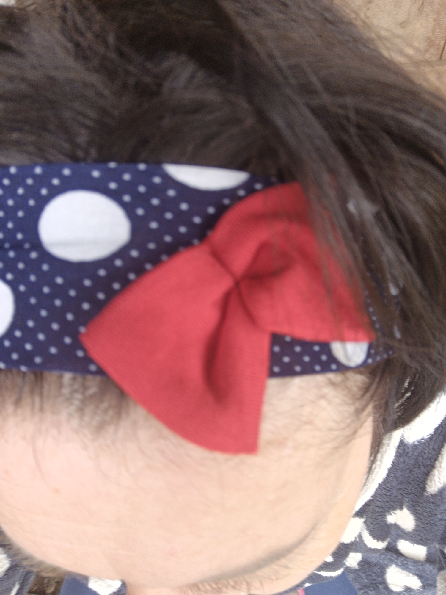 A close-up shot focuses on a person's head, specifically the forehead area. The person has dark hair, and a headband is worn across the forehead. The headband features a navy blue background with white polka dots and a red bow attached to the side. Part of the person's face, including an eye, is visible. A garment with a navy blue and white spotted pattern is also partially visible in the bottom right corner of the image.
No specific location in the city of Zalesovo, Russia can be determined from the image.