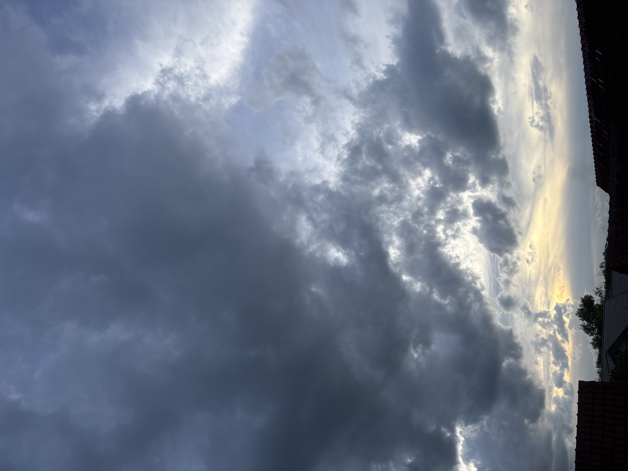 The image displays an expansive view of a heavily clouded sky over a residential area in บ้านบางรักษ์, Thailand, at what appears to be dusk or dawn. The upper two-thirds of the frame are dominated by dense, dark grey to bluish-grey cumuliform clouds, with the heaviest concentration on the left. Towards the right, the clouds are more fractured, revealing patches of lighter grey, pale blue, and white sky where light penetrates. A distinct yellowish-orange glow is visible in the lower sky, particularly towards the bottom-center and right, indicating the setting or rising sun behind the cloud cover. The bottom third of the image features the dark silhouettes of several rooftops. On the far left, a reddish-brown tiled roof is partially visible. In the bottom-center, a darker roof with a visible gable end is present, interspersed with the dark silhouettes of small trees. The far right side shows a horizontal segment of a dark, possibly corrugated, roofline. No people, animals, or specific actions are depicted.