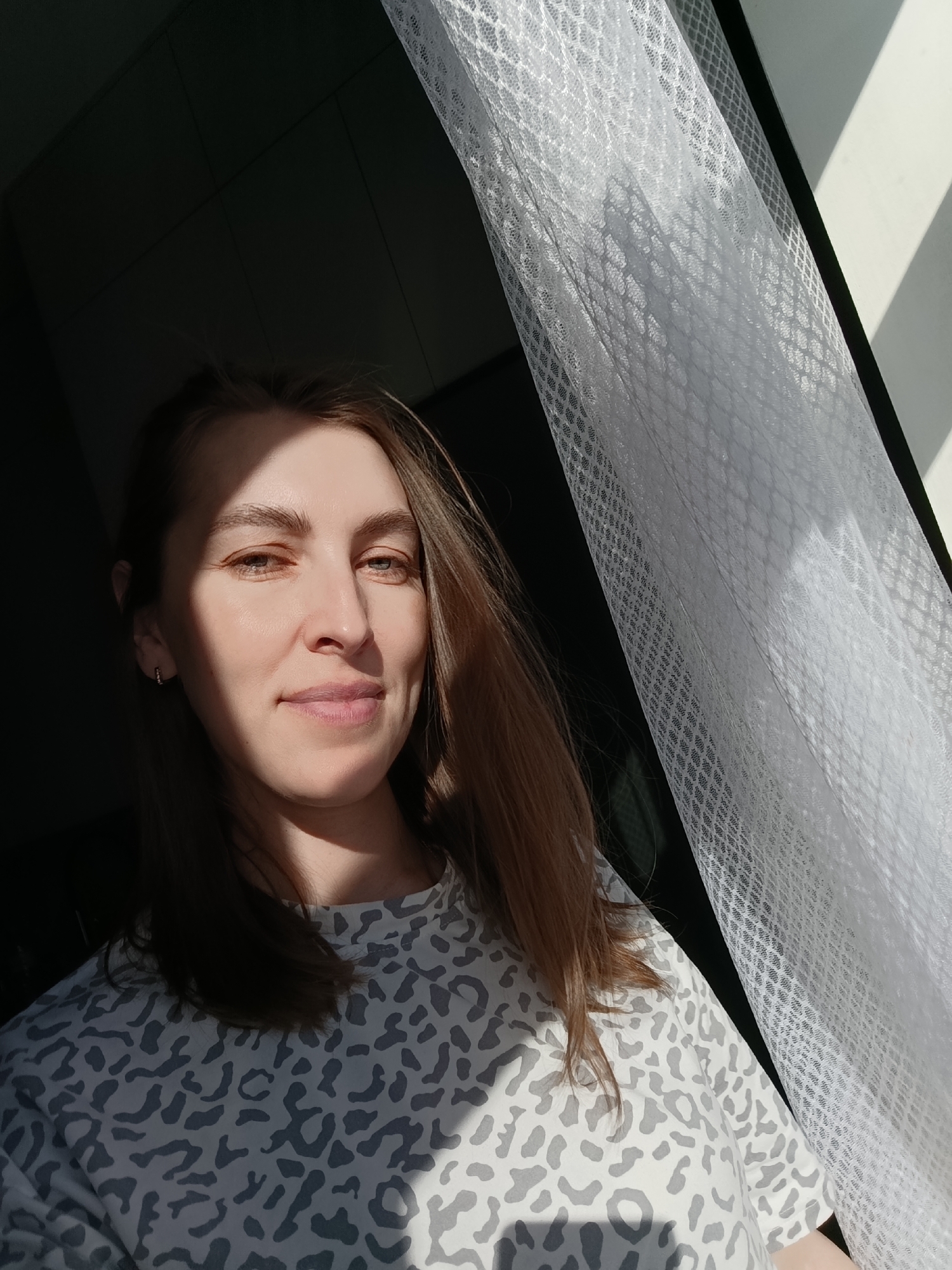 The image captures a woman indoors, likely in Chaikovsky, Russia, in what appears to be a selfie or a close-up portrait. She has long, straight brown hair, fair skin, and light-colored eyes, and is wearing a light-colored, long-sleeved top with a grey and white abstract or leopard print pattern. A small hoop earring is visible on her left ear.
The setting is an indoor space, with bright natural light coming from the right side of the frame, filtered through a sheer white curtain with a distinct honeycomb or mesh pattern. This strong sunlight creates dramatic light and shadow patterns on her face and shoulder, highlighting parts of her features while others remain in shadow. The background to the left is dark and obscured, possibly a wall or another part of the room in deep shadow.
The woman looks directly at the camera with a calm, serene expression, showing a subtle, gentle smile. The pronounced shadows and bright illumination suggest it is daytime, likely morning or late afternoon on a sunny day. There is no visible text in the image.