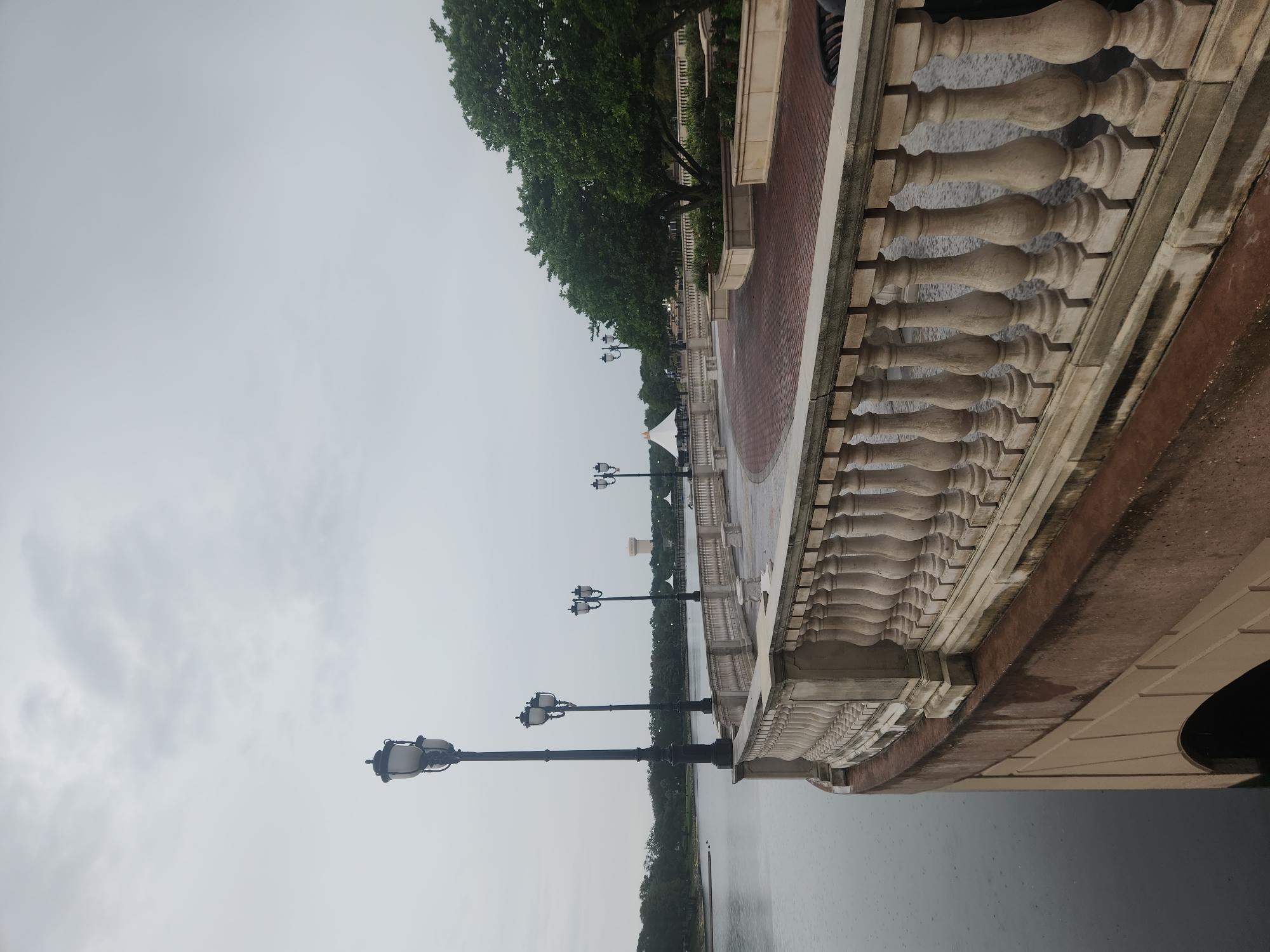 The image depicts a waterfront promenade with a ornate balustrade overlooking a calm body of water. The sky is overcast, suggesting a cloudy day. Several lampposts with traditional designs are visible along the pathway, which is paved with red bricks in a patterned arrangement. In the distance, a water tower and some structures, possibly gazebos or tents, can be seen. Trees line the far bank of the water, and the water itself shows ripples, indicating a light rain. The scene appears to be a public park or recreational area, likely in the city of Altamonte Springs, Florida, known for its lakes and parks. The architecture of the balustrade and lampposts suggests a somewhat formal or classical design aesthetic. No people are present in the image.