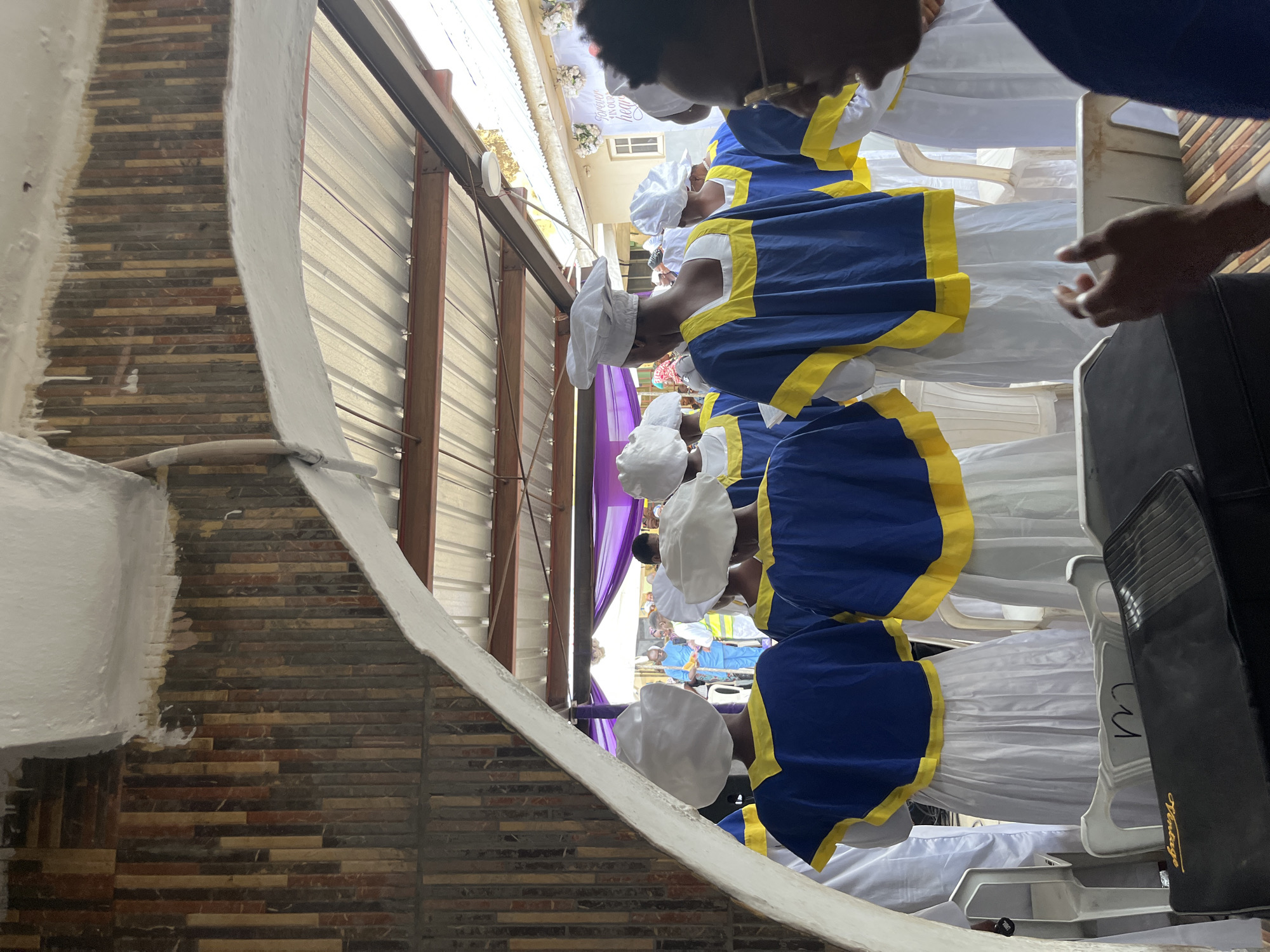 This media file depicts a group of people, likely women, dressed in matching white uniforms with blue and yellow trim, and white head coverings. They appear to be participating in a religious or ceremonial event. The individuals are lined up, facing away from the camera, and their attire suggests a formal occasion. The setting seems to be outdoors or in a covered outdoor space, with corrugated metal siding visible in the background. There are also structural elements like wooden beams and a white curved structure in the foreground, possibly a decorative element or part of a building.
In the background, a section of purple fabric is draped, adding a splash of color to the scene. Further back, glimpses of other individuals and more white and blue attire can be seen, indicating a larger gathering. The lighting suggests it might be daytime, possibly a bright day, but the specific time is not discernible. There are no clear indicators of weather conditions.
One of the cropped images shows a black case with the word "Vintage" printed in gold script. While the location is unknown, the overall atmosphere appears solemn and orderly, fitting for a procession or service. The focus is on the uniformity and presentation of the participants.