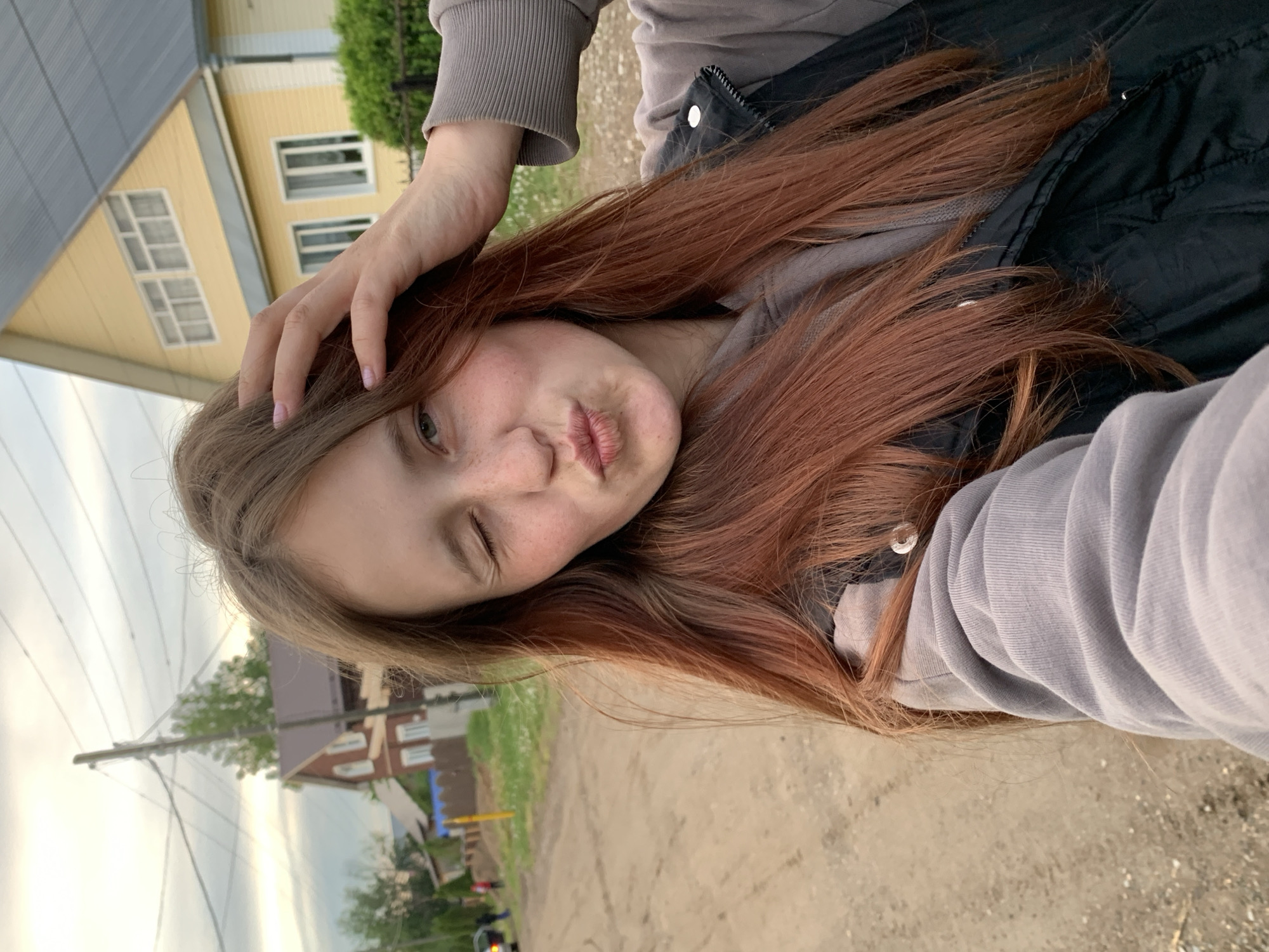 A young woman is taking a selfie outdoors. She has reddish-brown hair and is wearing a gray sweatshirt and a black vest. She is winking with her left eye and making a kissing face. Her right hand is touching her hair. The background shows houses and trees, suggesting a residential area. Based on the visible architecture and vegetation, the location is potentially Фоки, Russia. The ground appears to be a dirt or gravel road. The sky is overcast.