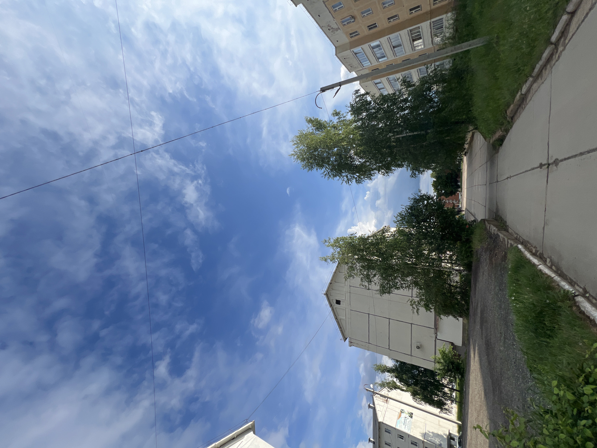 The image captures an upward-angled view of an urban street scene in Noviy, Russia. The sky, a dominant feature, is bright blue with scattered white clouds. Electrical wires stretch diagonally across the sky. On either side of the frame are buildings, typical of Soviet-era architecture. These buildings have a weathered appearance. Trees with green foliage are interspersed between the buildings and along the sidewalk. A paved walkway runs along one side of the image, bordered by a grassy area. The overall scene suggests a quiet, ordinary day in a residential neighborhood. No people are visible.