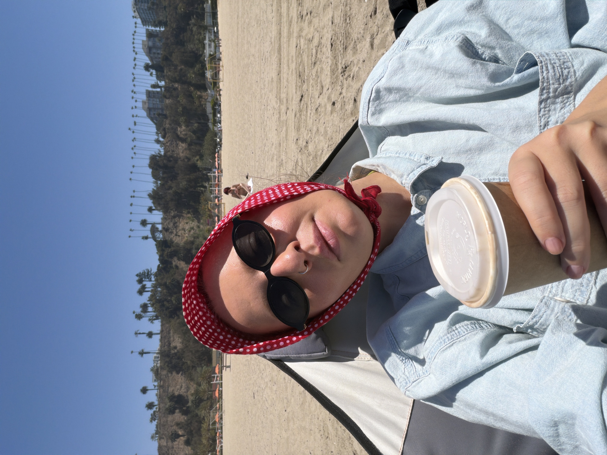 The image depicts a person resting at a beach in Santa Monica, United States of America. The person is fair-skinned and wearing a red and white polka-dot bandana, round sunglasses, and a light blue denim shirt. They have a nose ring and are holding a to-go coffee cup. The person is sitting in a beach chair with a gray and black frame. In the background, there is a sandy beach, palm trees, and buildings along the coastline. The sky is clear and blue, suggesting a sunny day.