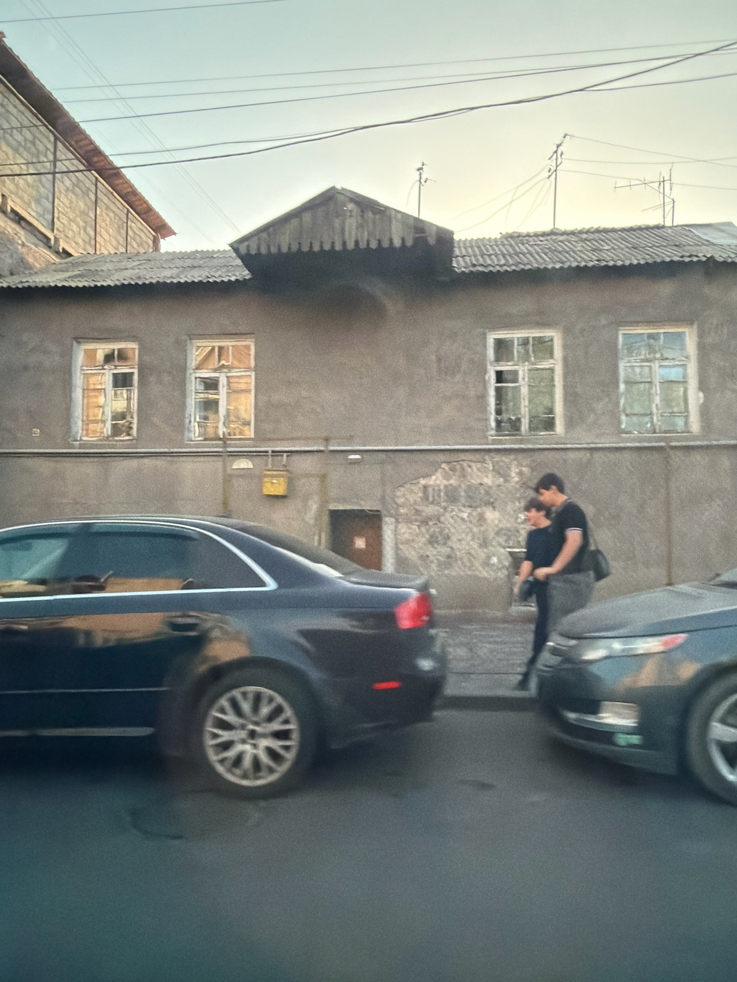 The image depicts a street scene in Yerevan, Armenia, with a focus on an aged, two-story residential building. The building's facade is a muted grey, showing signs of wear and tear, with several windows that appear to be in disrepair, some with broken panes and aged wooden frames. The roof is corrugated and appears weathered. Above the building, power lines crisscross the sky, with some antennas visible on the rooftop.
In the foreground, two cars are parked or moving on the asphalt street. A dark-colored sedan is prominently featured on the left, its metallic paint reflecting the ambient light. On the right, the front of a dark grey car is visible, its headlights and grille partially obscured.
On the sidewalk in front of the building, a couple is standing close together. The man, wearing a dark t-shirt and grey pants, has his arm around a woman who is wearing a dark top. They appear to be interacting, possibly in a romantic embrace. The background shows the rough texture of the building's wall and a small, yellow mailbox or utility box attached to the facade. The overall impression is of an urban environment with older architecture, set in the late afternoon or early evening given the lighting conditions.