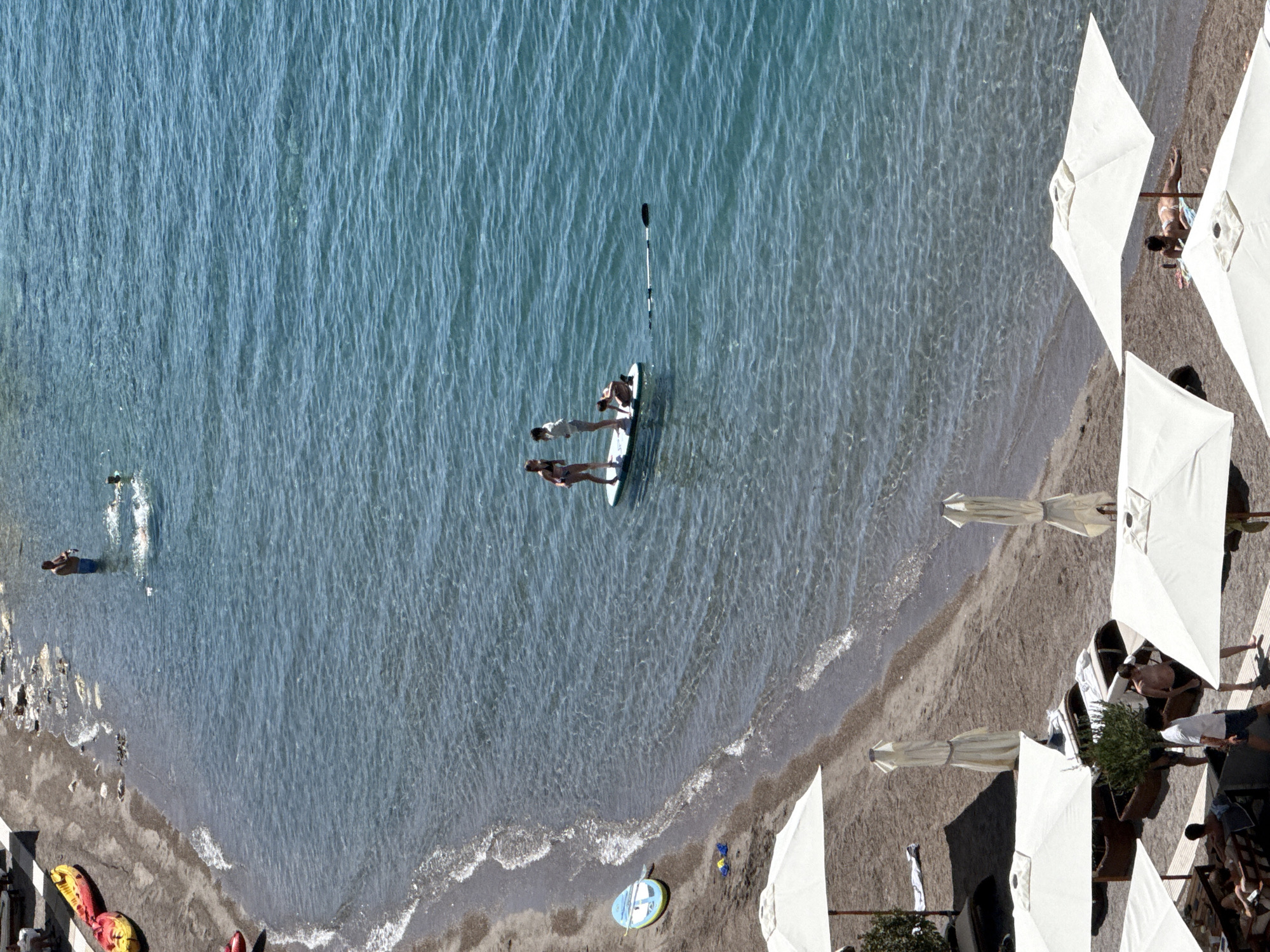 The image depicts a sunny day at a beach in Budva, Montenegro. The scene is divided between a sandy shoreline and clear blue water. On the beach, several white umbrellas are set up, providing shade for lounge chairs and a few people. Some individuals are seen relaxing on the beach or walking on wooden platforms.
In the shallow water near the shore, a person stands, and further out, two individuals are on a paddleboard. One person is standing, and another is crouching. A paddle lies beside the board. Further out in the water, another person is swimming.
On the sandy area of the beach, there are two yellow kayaks pulled up onto the shore. A blue and yellow paddleboard is also visible near the water's edge. The sea is calm with gentle waves lapping at the shore. The overall atmosphere suggests a leisurely day at a popular coastal resort.
