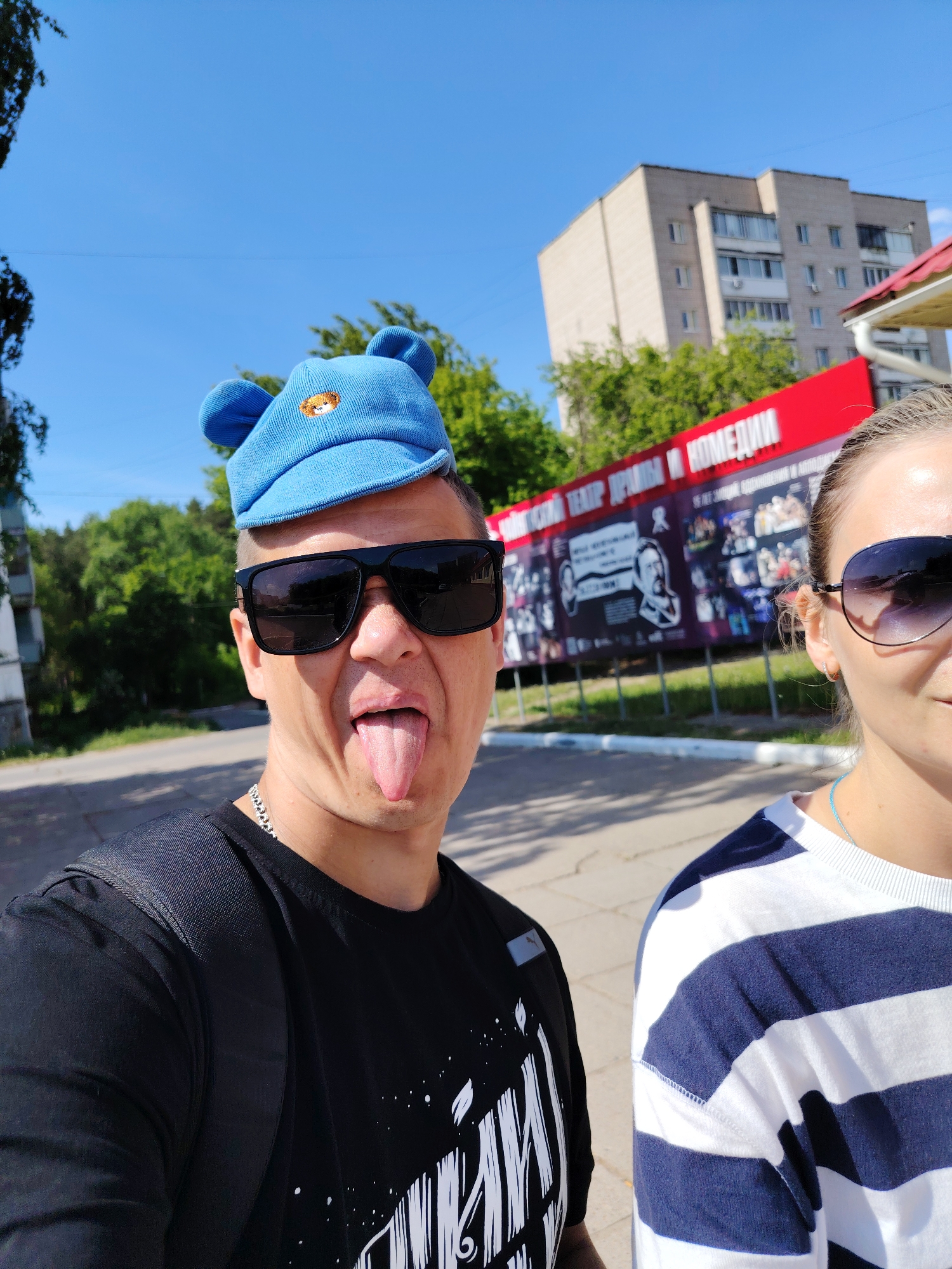 A medium shot captures a man and a woman taking a selfie outdoors in Chaikovsky, Russia. The man, positioned on the left, is wearing a black t-shirt, sunglasses, and a blue cap with ears. He is sticking his tongue out. The woman, on the right, is wearing a striped shirt and sunglasses. Behind them, a red sign is visible with a display of images and text in Russian. In the background, there is an apartment building, trees, and a blue sky. The scene appears to be taken on a sunny day.