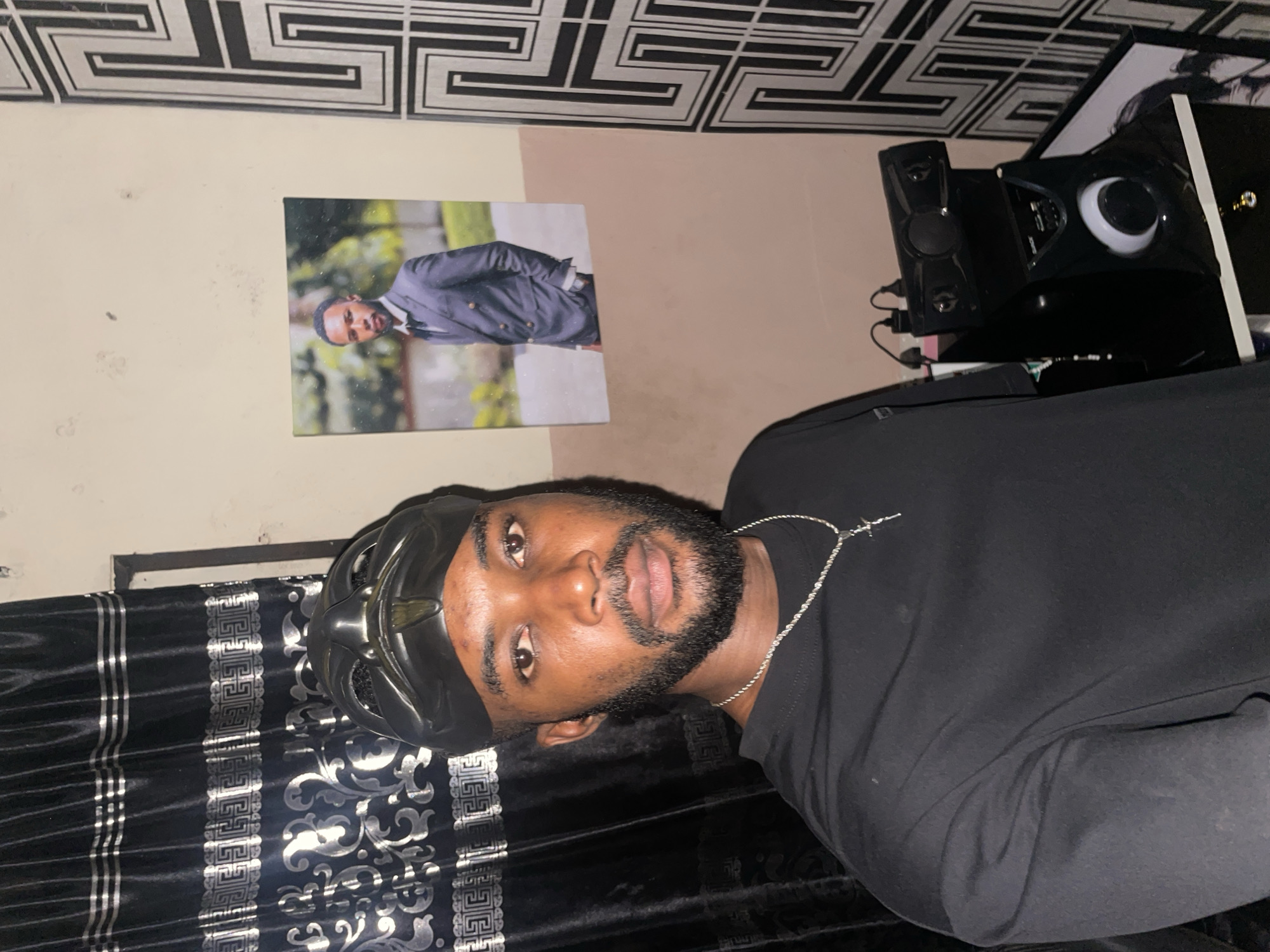 A man with dark hair and a beard is pictured indoors. He is wearing a black shirt and a black durag. A silver chain is around his neck. The background includes a wall with a picture on it, speakers, and decorative patterned wallpaper near the ceiling. It is not possible to determine the location in Lokoja, Nigeria, from the provided image.