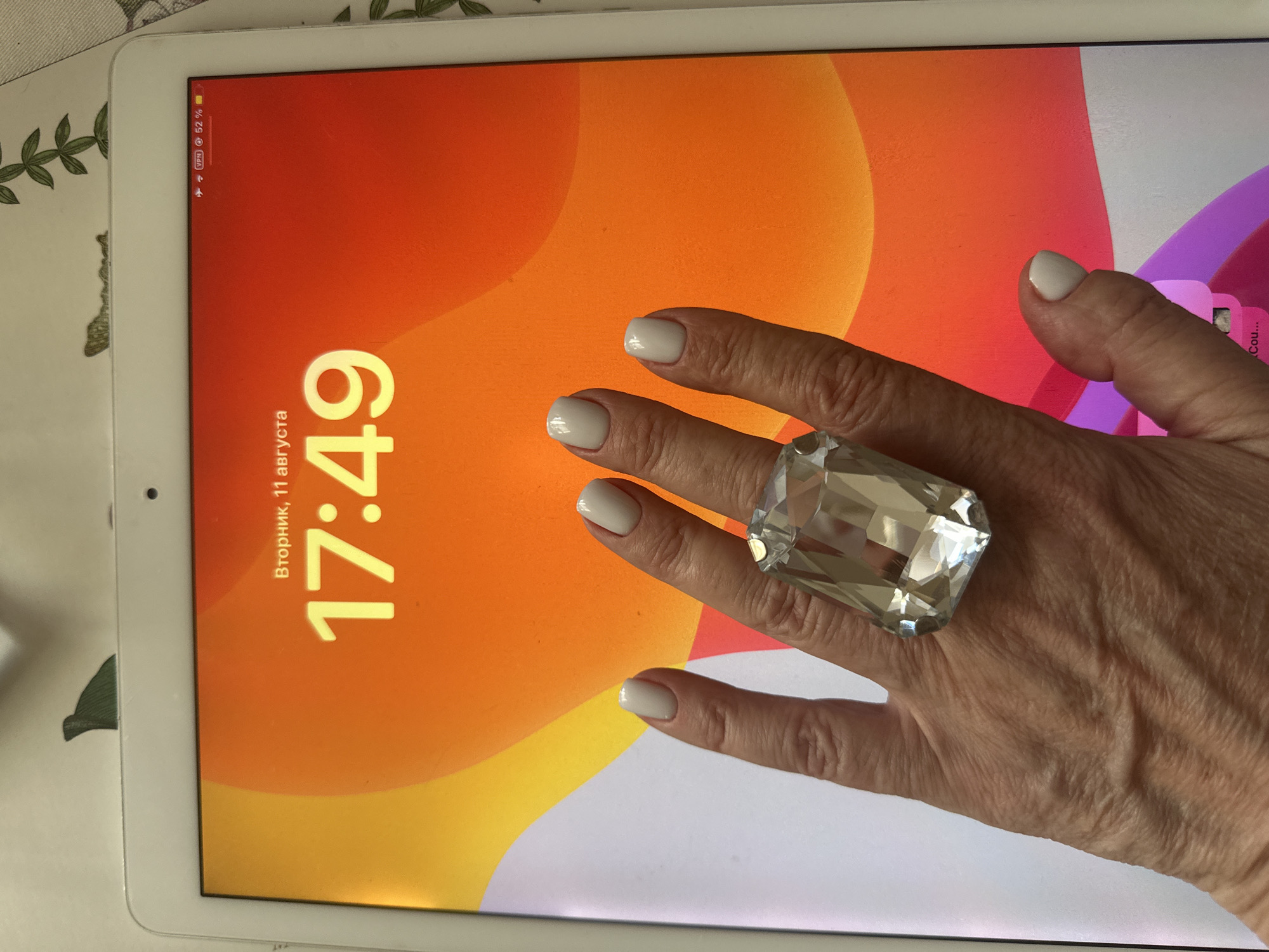 A close-up, eye-level shot shows a woman's hand with a large, clear, faceted gemstone ring on her index finger, superimposed over the lock screen of an iPad. The iPad displays the time as "17:49" and the date as "Вторник, 11 августа". The lock screen background is an abstract gradient of orange and yellow. The woman's nails are painted a light off-white color. The background of the image, outside of the iPad screen, is a lightly textured surface, possibly a tablecloth, with a subtle floral pattern. A small green object, possibly a leaf or a decorative item, is visible at the top edge of the frame. The image is well-lit, with the light source appearing to be from the top left. There are no discernible urban or environmental features that would indicate a specific city or location.