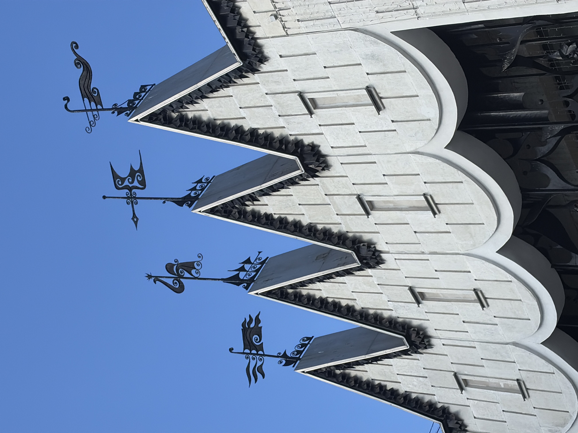 The image shows the facade of the Gorky Automobile Plant (GAZ) cultural center in Nizhny Novgorod, Russia. The building features a distinctive architectural style with a white, textured surface and a sawtooth roofline. Four black metal weather vanes are mounted on the peaks of the roof. The weather vanes are ornately designed with abstract shapes and stylized flags. The building has narrow, vertical windows with small metal lintels. The lower part of the facade features a series of arched decorative elements, below which is a dark, ornate metal grillwork that appears to be part of an entrance. The sky is clear and bright blue. There are no people visible in the image.
