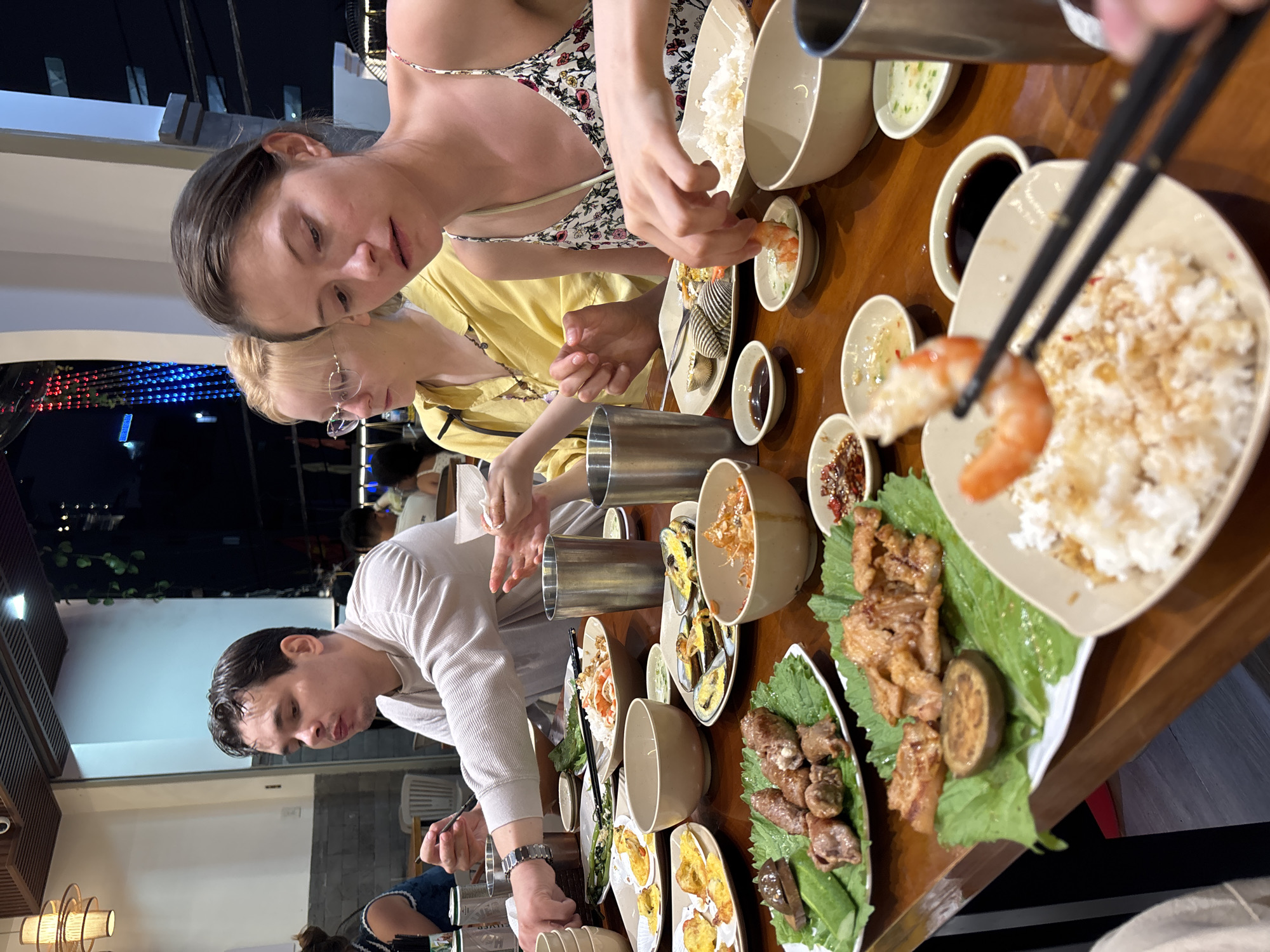 The image shows a group of people sitting at a wooden table, enjoying a meal. The scene is likely indoors, possibly a restaurant, with dim lighting suggesting it is evening or night. The people are engaged in eating and conversing, with a focus on the food laid out on the table.
Several individuals are visible. In the foreground, a man in a light-colored shirt is actively eating with chopsticks. To his left and further back, a woman with blonde hair and glasses is also seated at the table. The woman in the foreground, wearing a floral top, is reaching for food with her hand. Her expression is casual and engaged in the meal. The overall atmosphere appears relaxed and social, with people sharing a meal together.
The table is laden with various dishes, including bowls of rice, seafood like clams, and meat dishes. Small dishes of sauces and condiments are also present. Chopsticks and metal bowls are scattered across the table, indicating active dining. The background is blurred but shows what appears to be an indoor setting with some decorative elements, possibly lights.
The activity taking place is a meal shared among friends or family. The image captures a moment of shared enjoyment and camaraderie around food. There is no visible text in the image. The implied location is Nha Trang Ward, Vietnam, which would align with the type of cuisine and dining setting depicted. The time of day is likely evening, given the indoor lighting and the social context of a shared dinner.