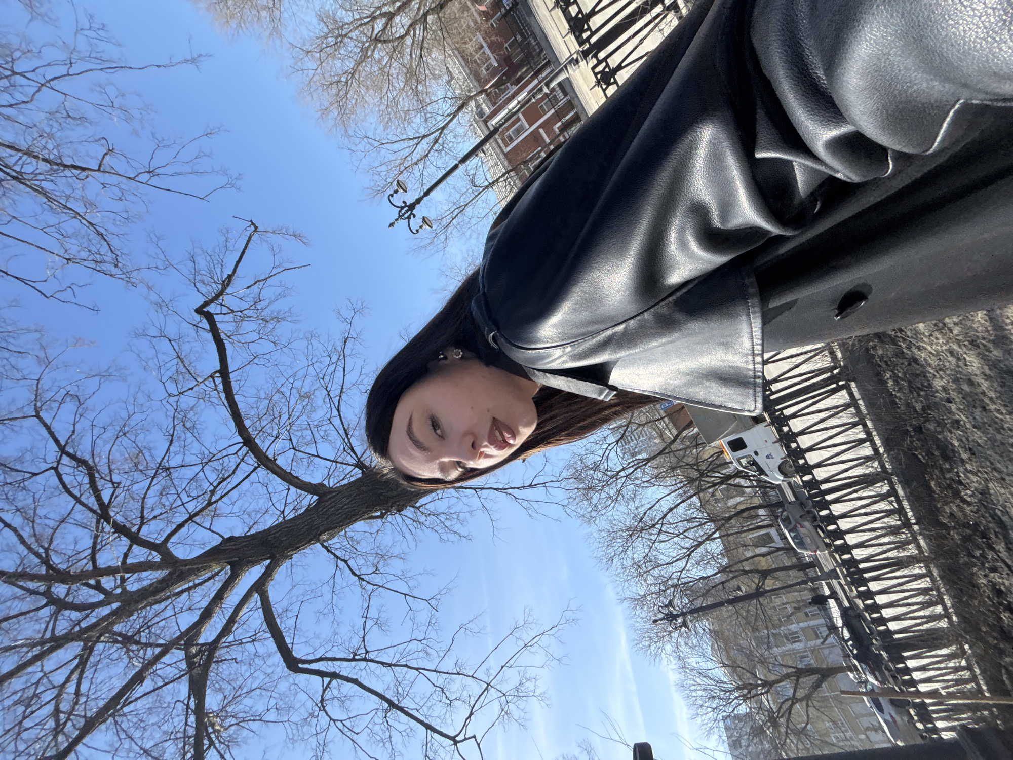 A woman in a black leather jacket stands under a tree with bare branches against a bright blue sky. A row of buildings with street lamps and parked cars is visible in the background, with a metal fence along the side. This scene is potentially located in the city of Perm, Russia.