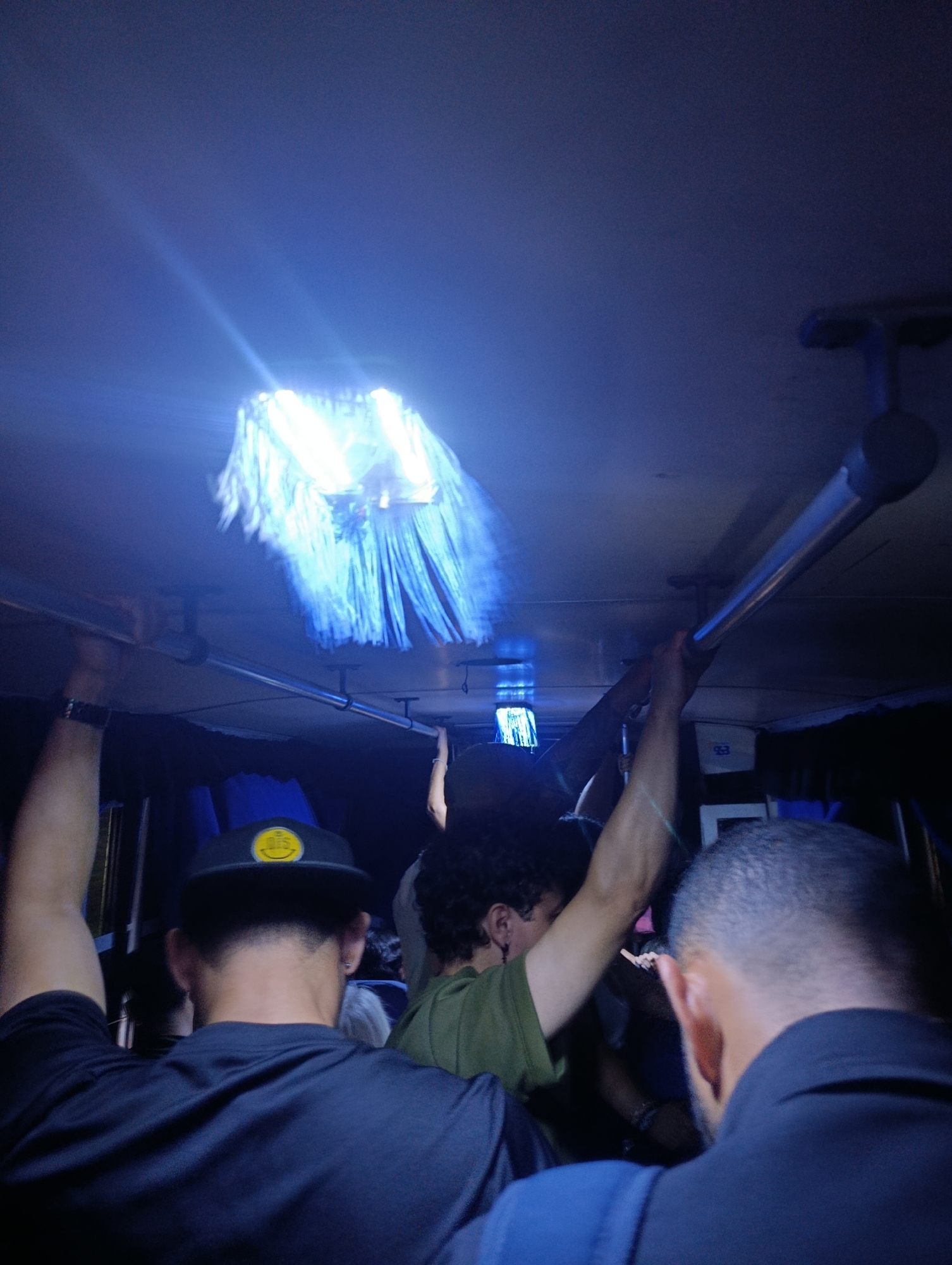 The photo depicts a group of people on a public bus in Juan Griego, Venezuela. The lighting is dim, illuminated primarily by a central overhead light fixture. Several passengers are visible, holding onto overhead bars for support. The scene conveys a sense of movement, possibly due to the bus being in transit. Visible passengers include one wearing a black t-shirt and cap, another wearing a green t-shirt, and another wearing a blue collared shirt.