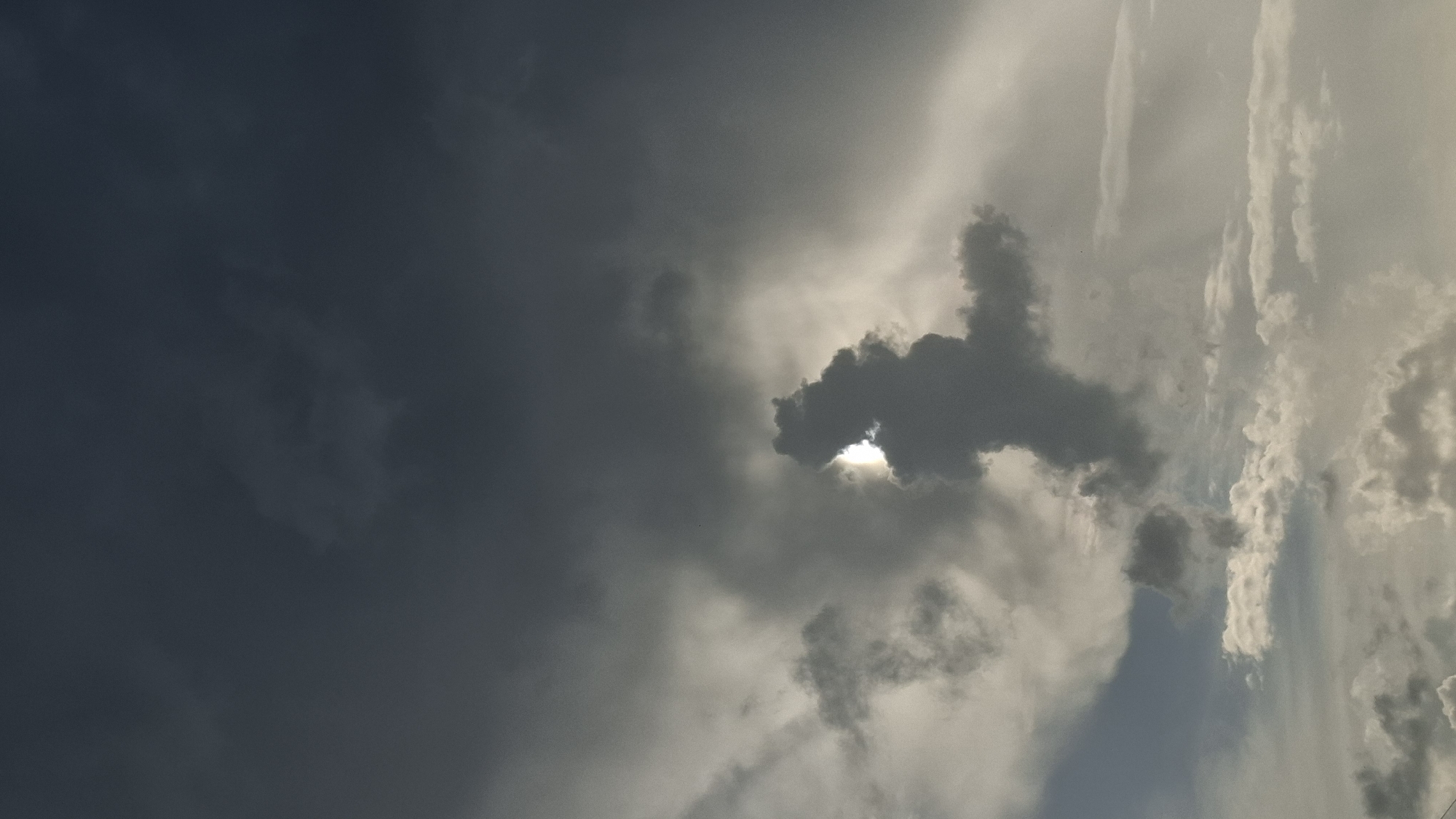 This is a photograph of the sky above Novaya Vasil'evka, Russia. The image is dominated by dark, dramatic clouds, with a sliver of bright sunlight peeking through a gap in the clouds. The light creates a halo effect around a dark cloud formation in the center of the frame that resembles a figure with outstretched arms. There are no people or man-made objects visible in the image. The overall mood is atmospheric and somewhat ominous, suggesting an approaching storm or the aftermath of one. The clouds are varied in texture and density, with some appearing wispy and others thick and imposing.