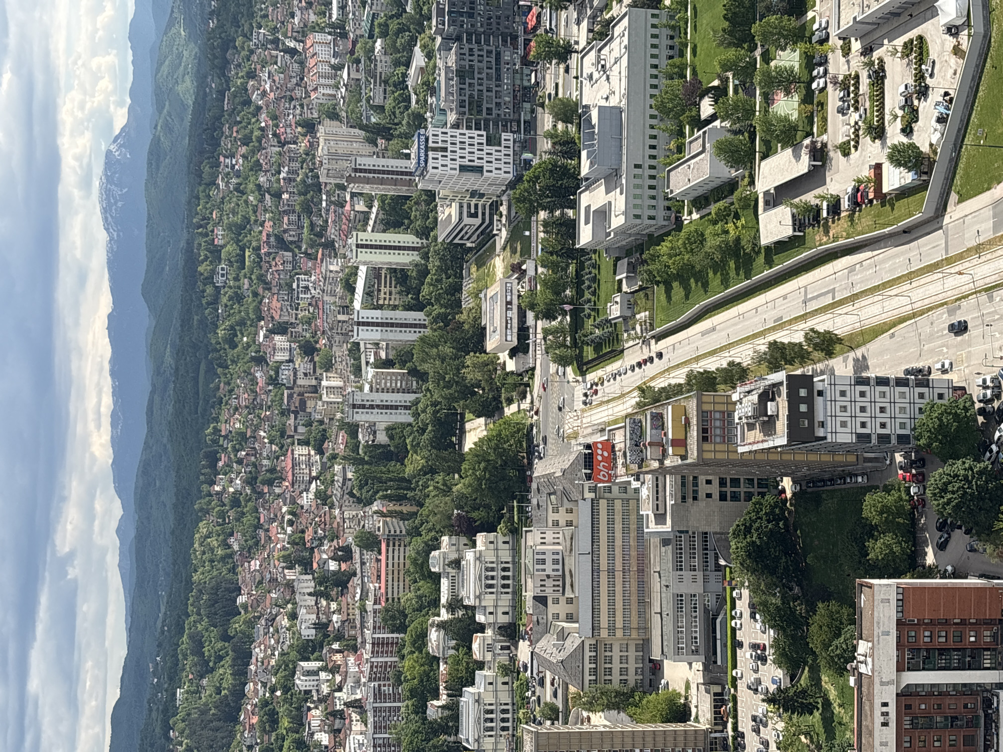 The image shows an aerial view of Sarajevo, Bosnia and Herzegovina, featuring a cityscape with a mix of residential and commercial buildings. Mountains are visible in the distance. A major thoroughfare runs through the city, with some vehicle traffic. Trees provide green spaces within the urban environment. The architecture varies, with a mix of older and modern buildings. Some parking lots and green areas are also visible.