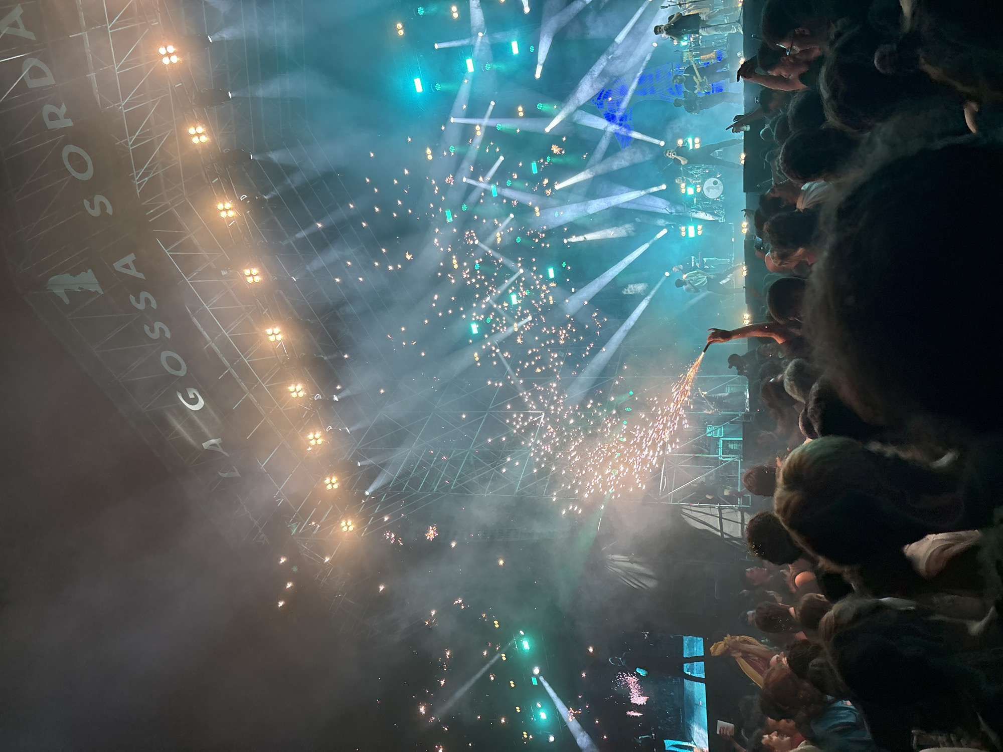 The image depicts a concert scene in Cunit, Spain, viewed from within the audience. A stage with lighting rigs is visible, along with performers. Fireworks are being set off, creating sparks and smoke. A crowd of people is present, some holding up their hands and recording with their devices. A sign reading "LA GOSSA SORDA" is visible. The environment appears to be a nighttime outdoor venue.