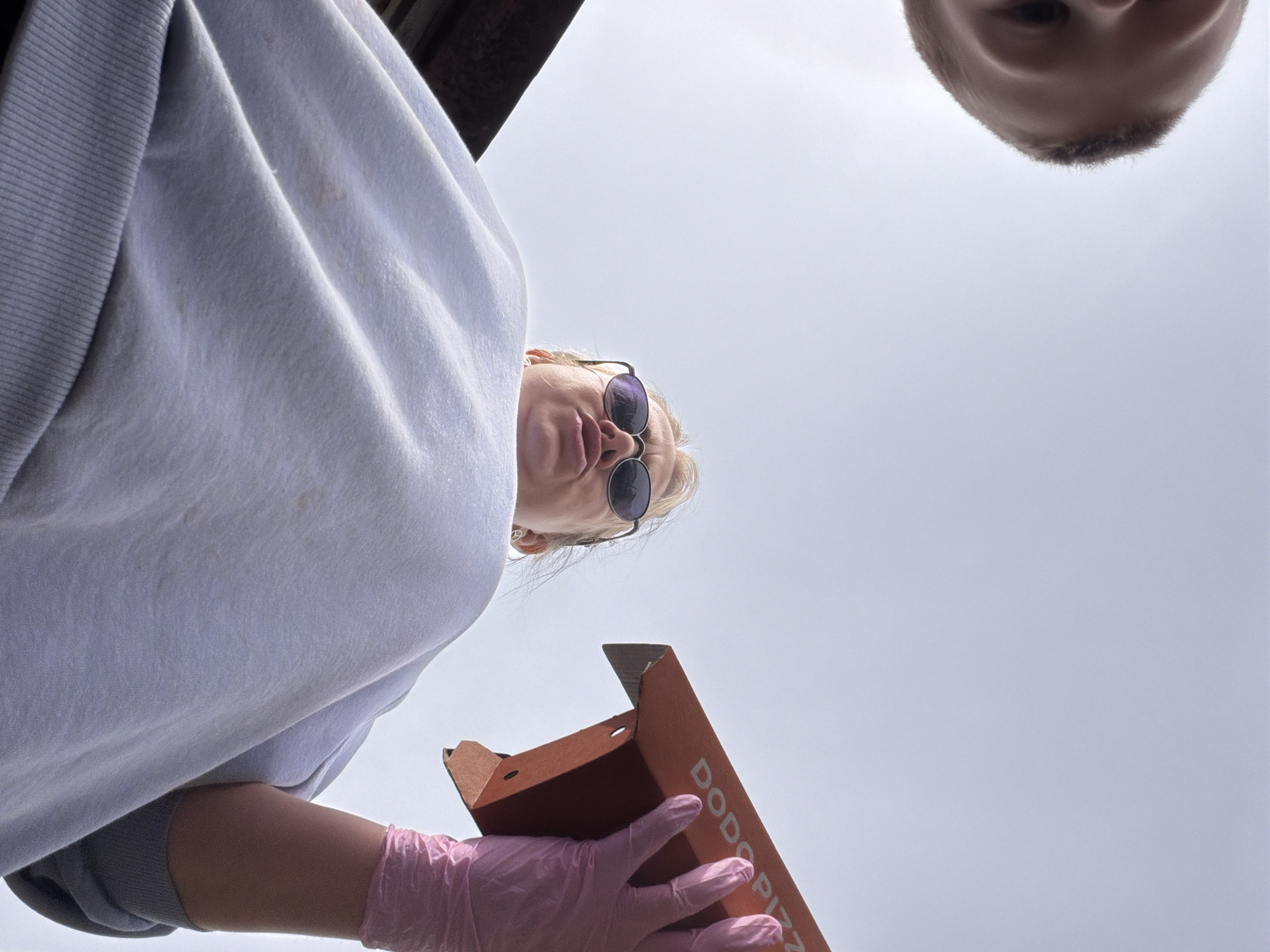 The image depicts two individuals and a pizza box from a low-angle perspective against a gray sky. A woman with blonde hair and sunglasses is visible, wearing a white sweatshirt and pink gloves. She holds an open brown pizza box with the words "DODO PIZZA" printed on it. Above her, partially visible, is the head of a child with short, light-colored hair. The location is Бронница, Russia, indicated by the "DODO PIZZA" box, as this company operates in that city.