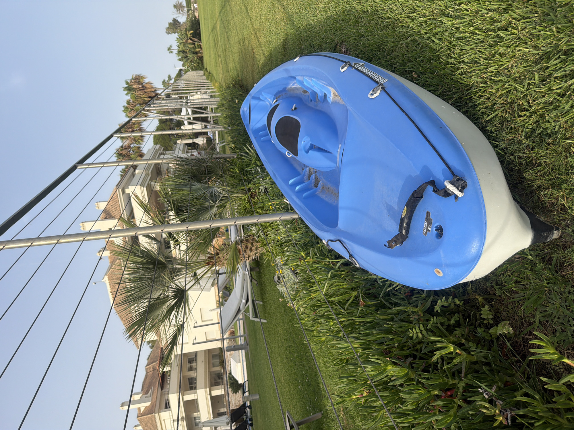 A blue and white kayak rests on a grassy area next to a metal fence. The kayak is oriented diagonally, with its front pointing towards the bottom left of the frame. The background shows a building with a tiled roof, palm trees, and lounge chairs, suggesting a resort or residential area in a warm climate. The sky is clear and blue. The scene appears to be in Estepona, Spain, based on the architectural style and vegetation.