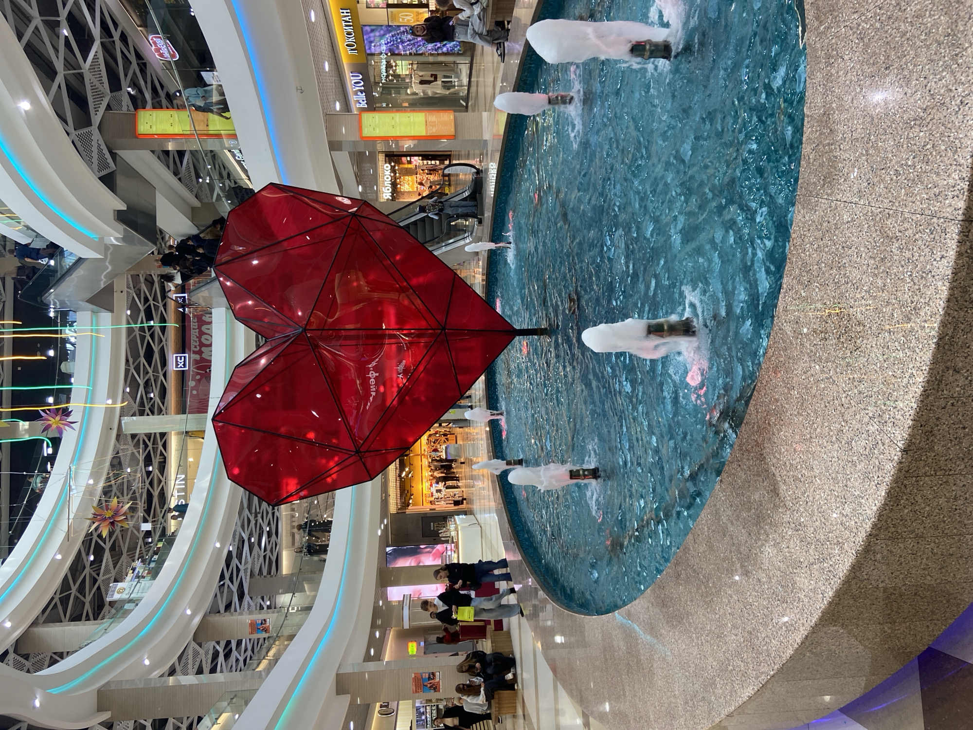 The image shows the interior of a modern shopping mall in Irkutsk, Russia. In the foreground, a circular fountain with bright blue water is visible, with several water jets creating frothy plumes. A large, geometric red heart sculpture is positioned in the center of the fountain. The mall features multiple levels with balconies and escalators, illuminated by colorful LED lighting strips. Several people are seen in the background, some sitting on benches, others walking, and some on escalators. Shops with illuminated signs are visible on different floors. The architecture is contemporary, with a focus on bright and open spaces.