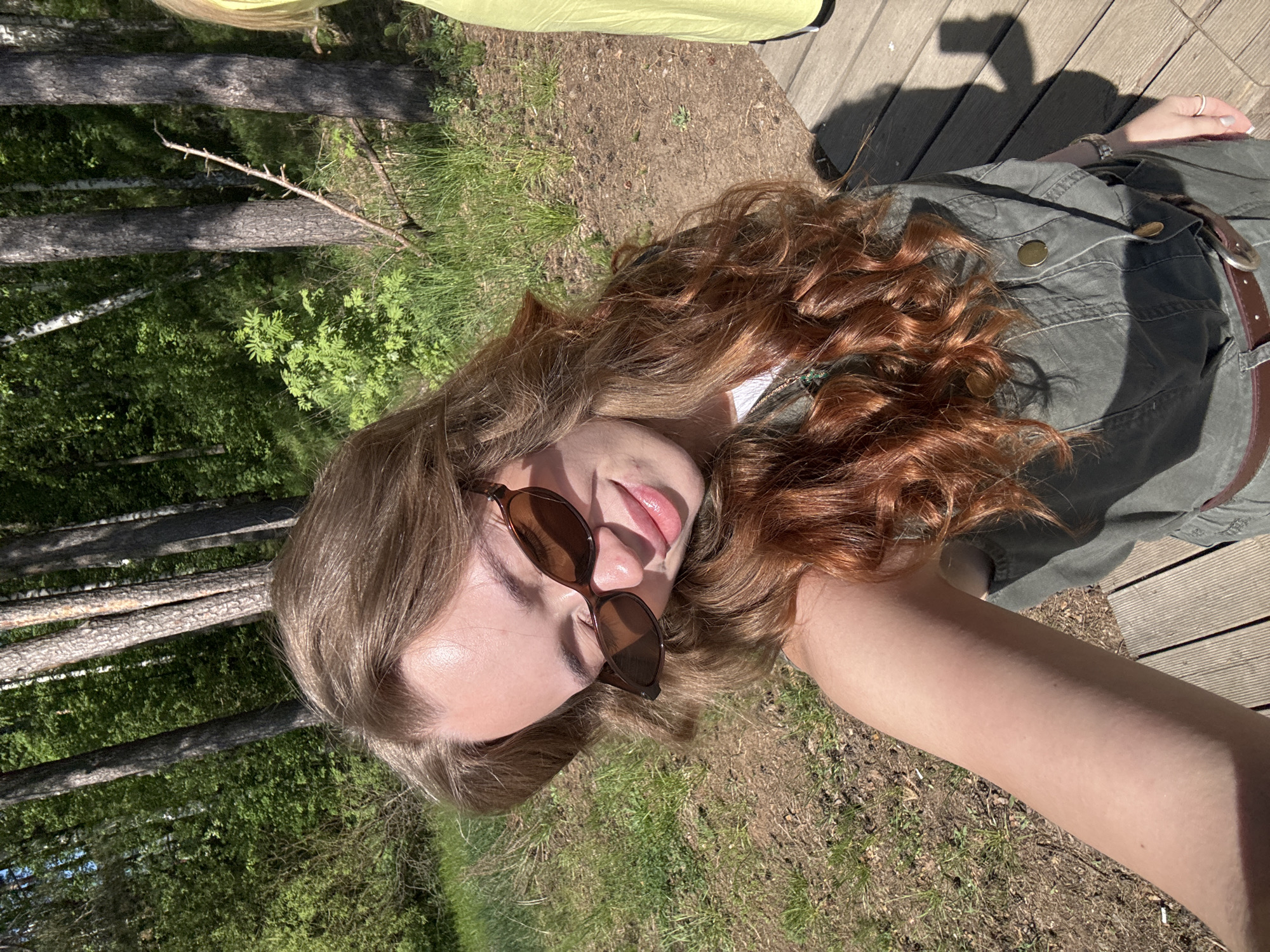 A young woman poses for a selfie outdoors. She is wearing sunglasses and a green jacket with a brown belt. Her hair is light brown with auburn highlights. She is positioned on what appears to be a wooden deck or platform. In the background, there are trees and grassy areas, suggesting a park or wooded area. A patch of yellow fabric is also visible, suggesting another person nearby. It is daytime, based on the lighting and shadows. The exact location in Chaikovsky, Russia cannot be determined based on the provided information.