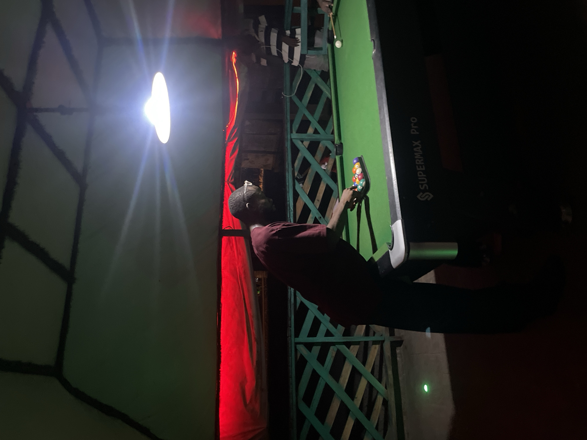 A full-shot photograph, taken at night, depicts a group of people playing pool at an outdoor venue in Lokoja, Nigeria. The scene is illuminated by a bright overhead light and ambient red lighting from above. In the foreground, a person wearing a maroon t-shirt and glasses is bent over a green pool table, reaching for a rack of colorful pool balls. To the right of this person, another individual in a striped shirt stands near the pool table. The environment includes a wooden railing with a green patterned design. The pool table itself has the "SUPERMAX Pro" logo visible on its side. A small green light is also visible on the ground to the left of the pool table.