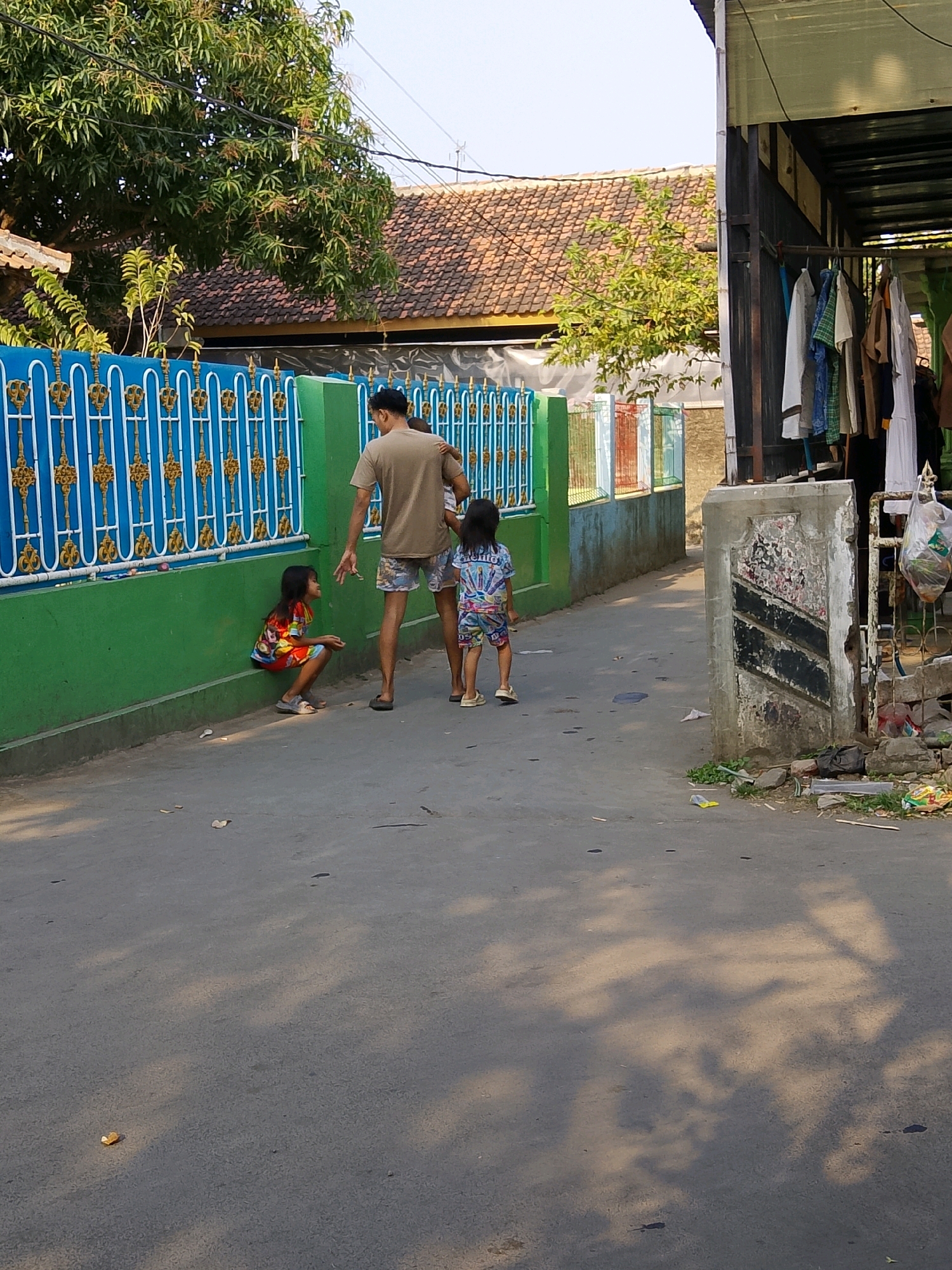 The image depicts a residential alleyway in Cirebon, Indonesia. The scene features a man in a tan t-shirt and patterned shorts interacting with two young girls. One girl, wearing a colorful dress, is crouched down near a green wall with a blue and gold patterned fence. The other girl, in a t-shirt and shorts with a similar pattern, stands further down the alley. To the right, a covered structure with clothes hanging on a rack is visible, along with piles of debris and construction materials at the base of a weathered concrete pillar. The alleyway itself is paved, and shadows from overhead trees are cast across the surface. The background includes a house with a tiled roof and lush green trees. The environment suggests a warm, sunny day in a densely populated urban or suburban area.
