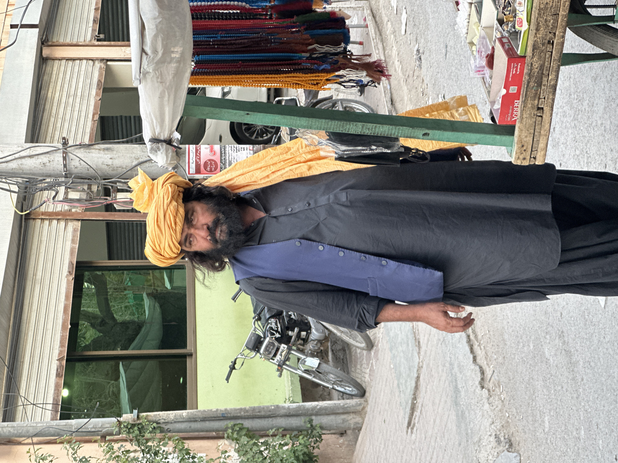 The image captures a man, presumably in Quetta City Tehsil, Pakistan. He has a turban on his head, is wearing a black kurta and a blue vest. He is standing on a concrete pavement in front of a building with closed rolling shutters. A motorcycle is parked nearby. A vendor's cart is visible with merchandise on it. The cart includes prayer beads of various colors, packaged goods. Signs with text in Urdu are also visible, including one with the word "BERRA" and another that has "Knitting Caps" on it. Wires are tangled overhead. Green plants are seen.