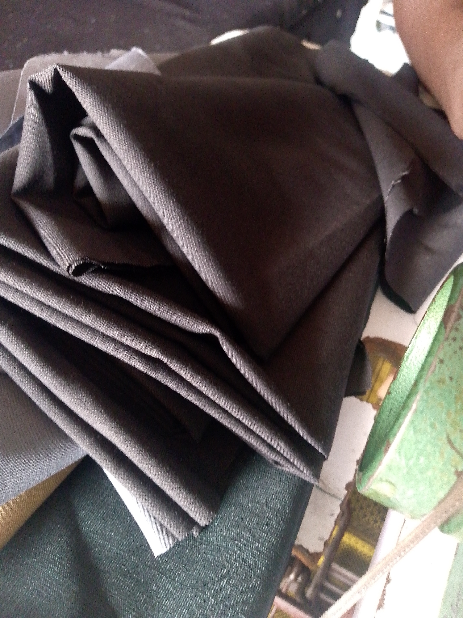 The image shows a pile of folded dark-colored fabric. The fabric appears to be a synthetic blend, possibly polyester or a polyester-cotton blend, given its smooth texture and lack of prominent weave. The folds are sharp, suggesting the fabric is somewhat stiff and has been pressed or is new. In the background, a green object, possibly a bowl or container, is partially visible, along with some indeterminate blurry shapes. The lighting is dim, casting shadows on the fabric, which contributes to the dark overall tone of the image. There are no visible people or explicit signs of a specific location within Jauro Yinu, Nigeria. The scene is static and focuses solely on the fabric and its immediate surroundings.