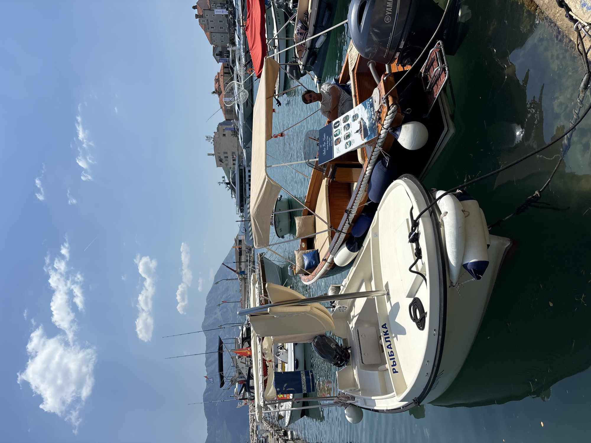 This is a vertical, eye-level shot of a marina with several boats docked. In the foreground, a white boat with blue trim is visible, with the Cyrillic word "РЫБАЛКА" (Rybalka) emblazoned on its stern. It has a small outboard motor and fishing rods are attached to its sides. Behind this boat, a wooden boat with a beige canopy is docked. A person wearing sunglasses and a striped shirt is sitting in this wooden boat, looking towards the camera. A menu board with pictures of boats is also visible on the wooden boat.
In the background, more boats are docked, and beyond them, buildings with terracotta roofs are visible, suggesting a coastal town. Mountains rise in the distance, some of which are covered in greenery. The sky is bright blue with scattered white clouds, indicating a sunny day, likely late morning or early afternoon. The water is calm and a deep green, reflecting the boats and the sky. The overall scene depicts a peaceful day at a marina, possibly for leisure boating or fishing.