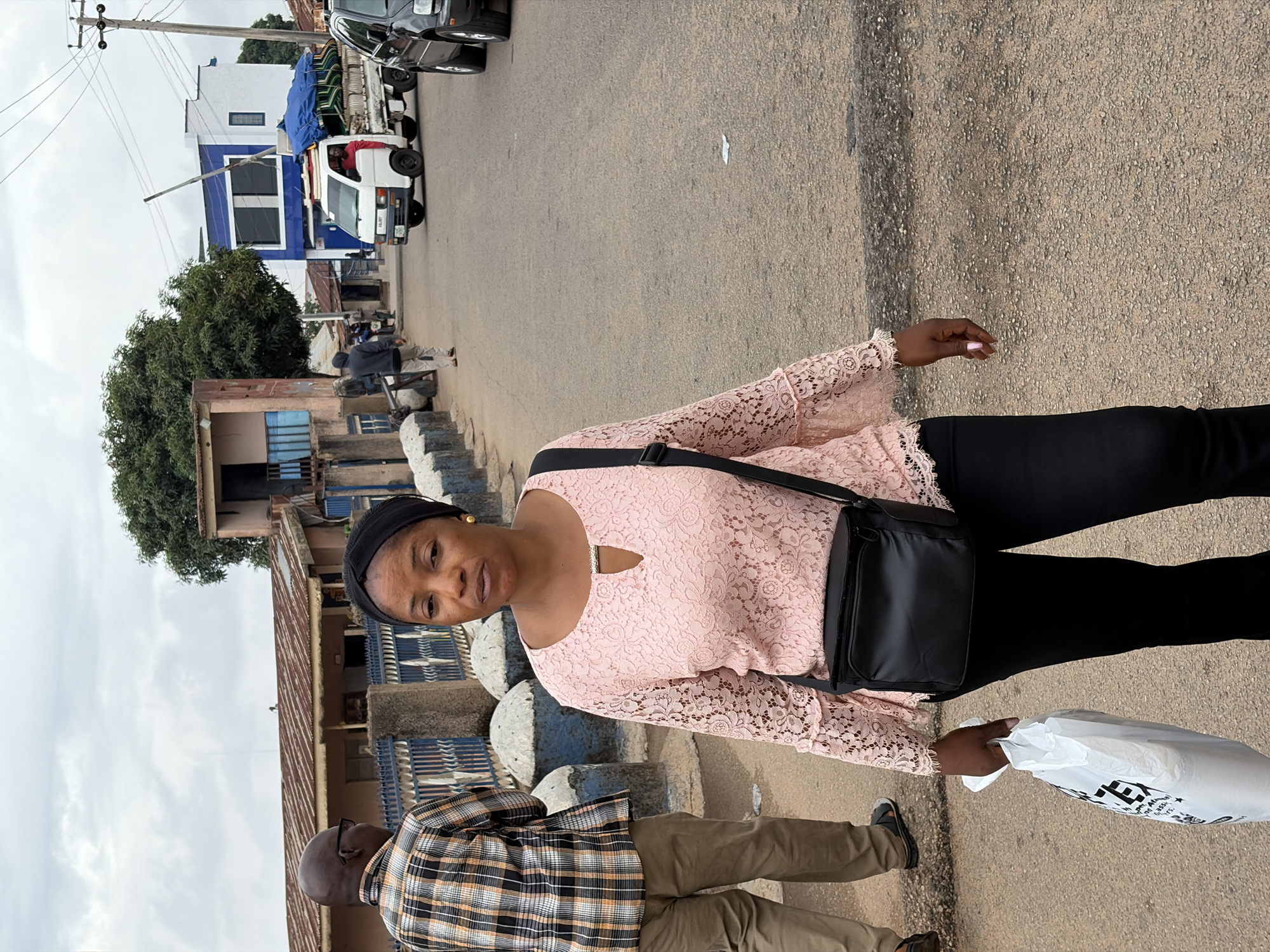 A woman wearing a pink lace top and black pants stands on a paved street in Jos, Nigeria. She has a black head covering and a black purse is slung over her shoulder. She holds a white plastic bag in her left hand. A man in a plaid shirt and khaki pants walks to her right. A building with a blue and white facade is visible in the background, along with a white pickup truck. Power lines stretch across the top of the frame.