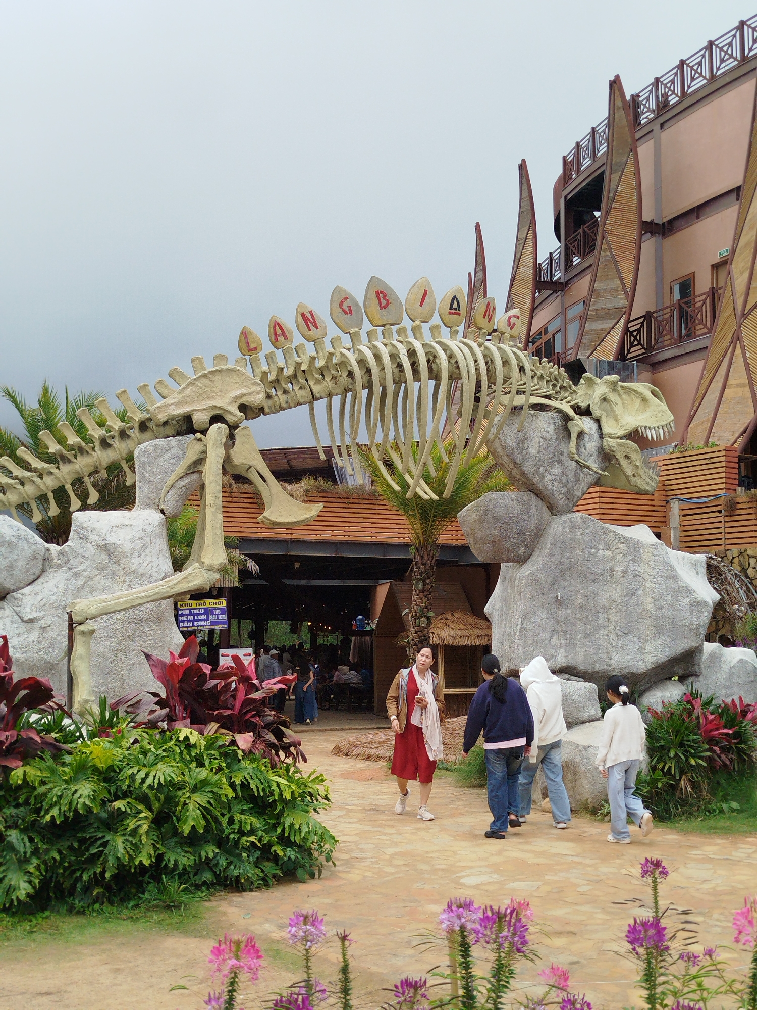 This image shows the exterior of the Lạc Dương Dinosaur Park in Lạc Dương, Vietnam. A large, beige dinosaur skeleton, likely a Stegosaurus based on the plates along its spine, is prominently displayed. The skeleton is propped up by large grey rocks. The word "LANGBA" is visible in red letters on the dinosaur's plates. In the background, a multi-story building with a unique architectural style featuring wooden beams and a salmon-colored facade stands. Several people are walking in the foreground and midground. On the left, a woman in a red dress and beige scarf is walking away from the viewer. To her right, three individuals, two in hoodies and jeans and one in a white sweater and jeans, are walking towards the building. Further right, a young girl in a white sweater and light blue jeans is walking away from the viewer. Lush green foliage and red-leafed plants are visible around the base of the dinosaur skeleton. Pink and purple flowers are in the foreground. The ground is a mix of dirt and paved walkway. The sky is overcast.