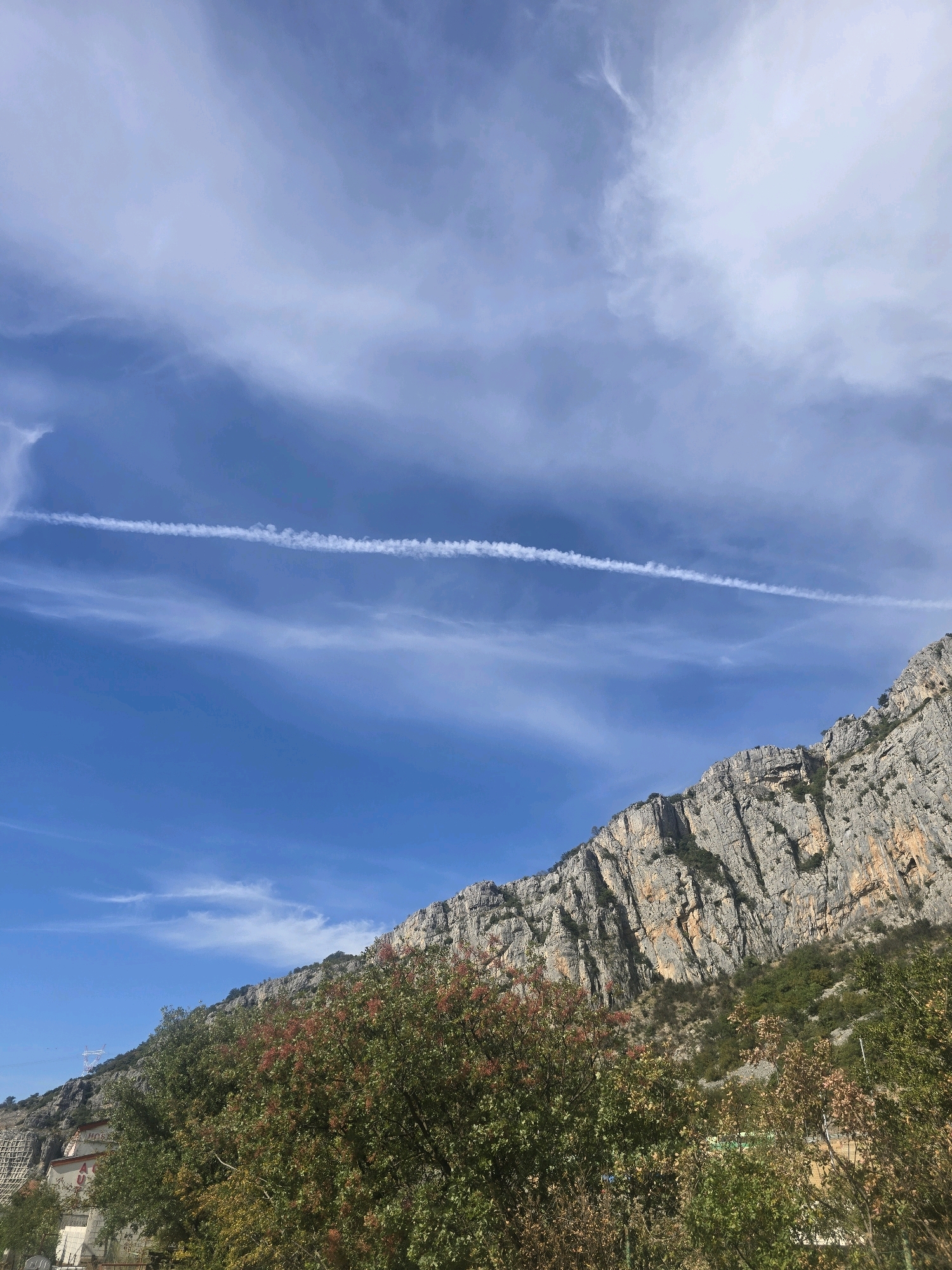 The image depicts a mountainous landscape under a bright blue sky with wispy clouds and a prominent contrail from an aircraft streaking across the upper portion. In the foreground, lush green and reddish foliage is visible, indicating the presence of trees and bushes. Beyond the vegetation, a steep, rocky mountainside dominates the right side of the frame, with visible textures and geological formations. The lower left corner of the image shows a glimpse of buildings and infrastructure, suggesting an urban or semi-urban environment at the base of the mountain. This scene is located in the city of Podgorica, Montenegro, a region characterized by its proximity to mountainous terrain and a significant river.