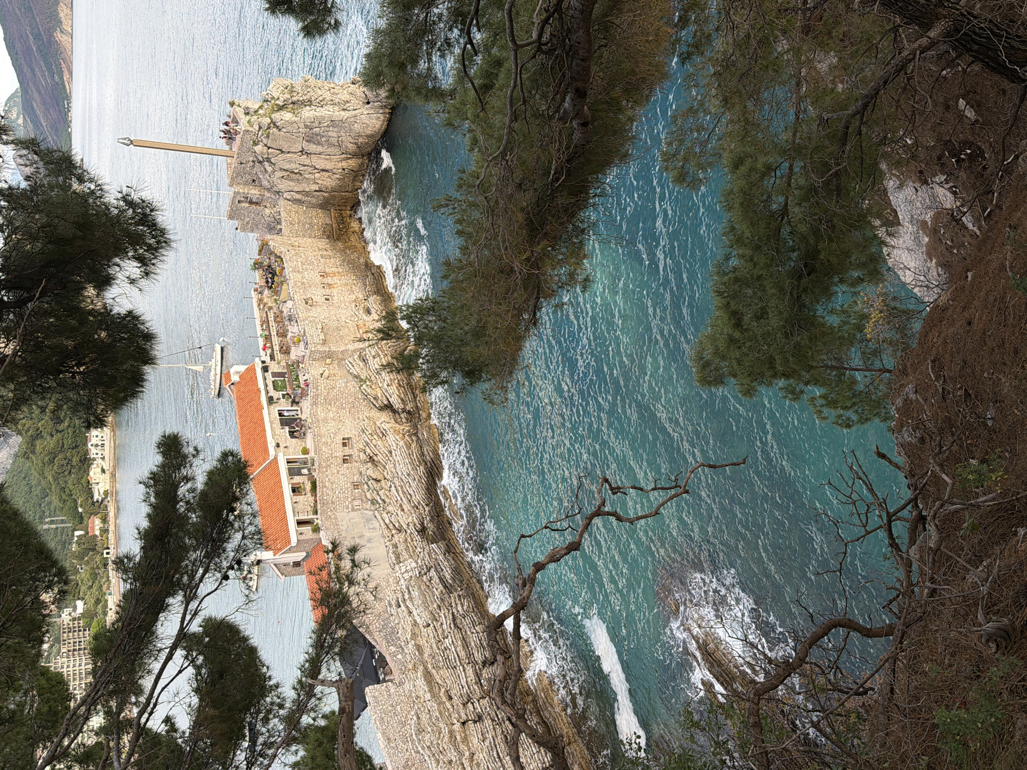 A high-angle, outdoor view depicts a coastal scene in Petrovac centar, Montenegro, showing a stone fortress and surrounding landscape. A structure with a red tile roof is visible on the left, partially obscured by trees. A stone wall extends from this structure towards the ocean.
A rocky promontory extends into the turquoise water, with trees hanging over the edge. People are present on top of the rocky promontory. The ocean surface is textured by ripples and whitecaps, with the shoreline marked by crashing waves.
Trees frame the view from the foreground, obscuring parts of the scene. The distant land across the water is faintly visible.
