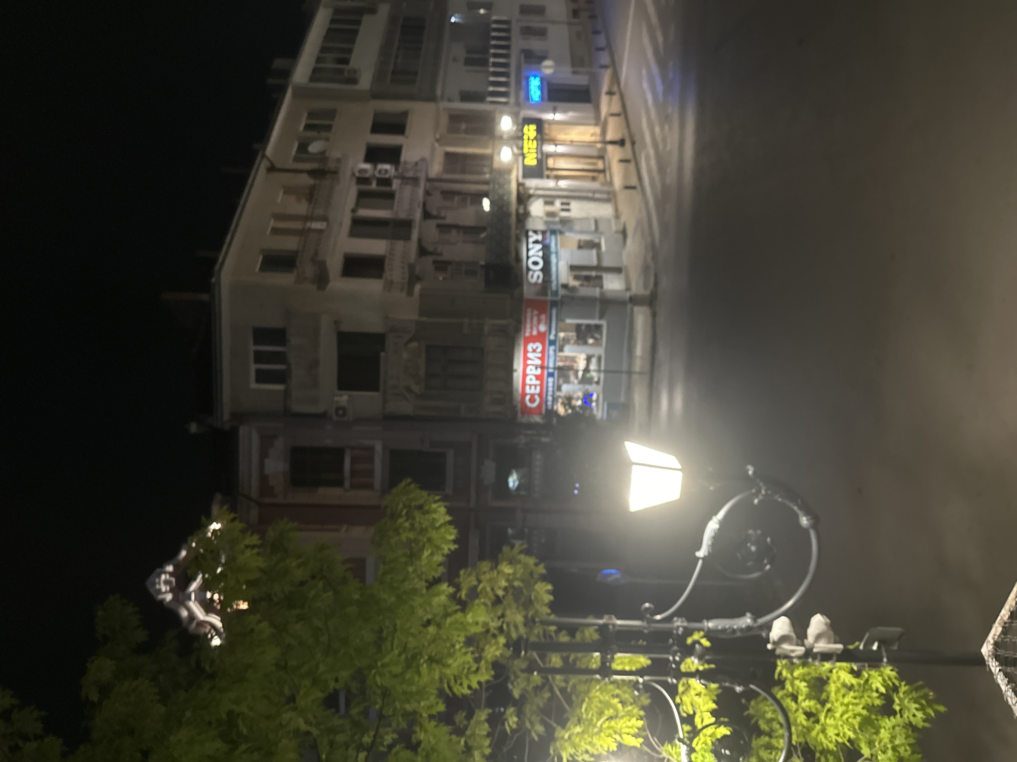 The image depicts a nighttime street scene in Ruse, Bulgaria, at the intersection of Tsar Ivan Shishman Street and General Skobelev Street. The scene is dominated by the architecture of the city, with several multi-story buildings lining the streets. On the left side of the frame, a vibrant green tree partially obscures the view of a building with an ornate, illuminated rooftop. In the foreground, a streetlamp with an illuminated, square-shaped light fixture casts a warm glow on the surrounding foliage and the dark street. The street itself appears to be wet, reflecting the lights from the buildings.
Several businesses are visible along the street. One building features prominent signage for "SERIZ" and "SONY," indicating retail establishments. Another building displays signage that reads "INTERA." The street is mostly devoid of people, suggesting a quiet late-night or early-morning atmosphere. Pedestrian crossings are visible on the asphalt, and bollards line the sidewalk in front of some of the businesses. The overall ambiance is that of a tranquil, urban environment at night.