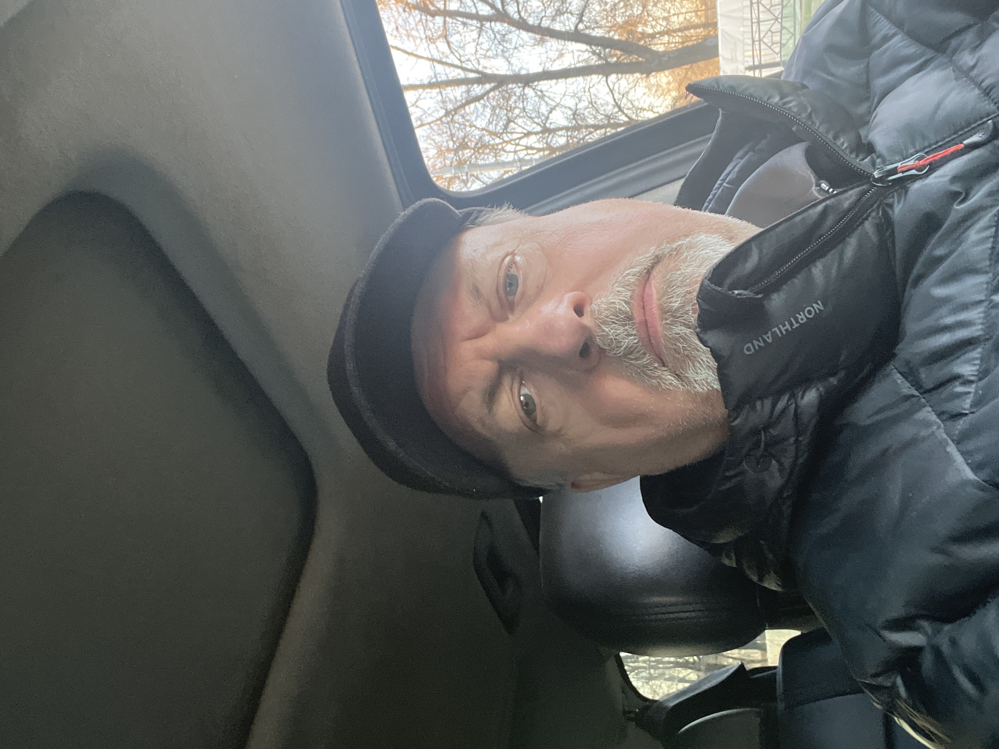 A man is pictured from a low angle inside a vehicle, likely a car, with the roof open, partially revealing bare tree branches against a bright sky. He is wearing a dark-colored, puffy jacket with the word "NORTHLAND" visible on the collar and a dark cap. The man has fair skin, short gray facial hair, and appears to be looking toward the camera. The interior of the vehicle is gray and includes a headrest. The location, Podolsk, Russia, cannot be confirmed from the image.