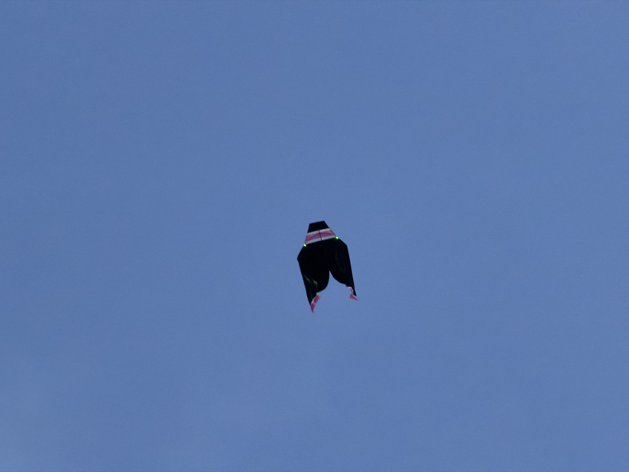 A black, pants-shaped kite with pink accents flies against a clear blue sky. The location is unknown, and no information indicates that it is in North Kuta, Indonesia.