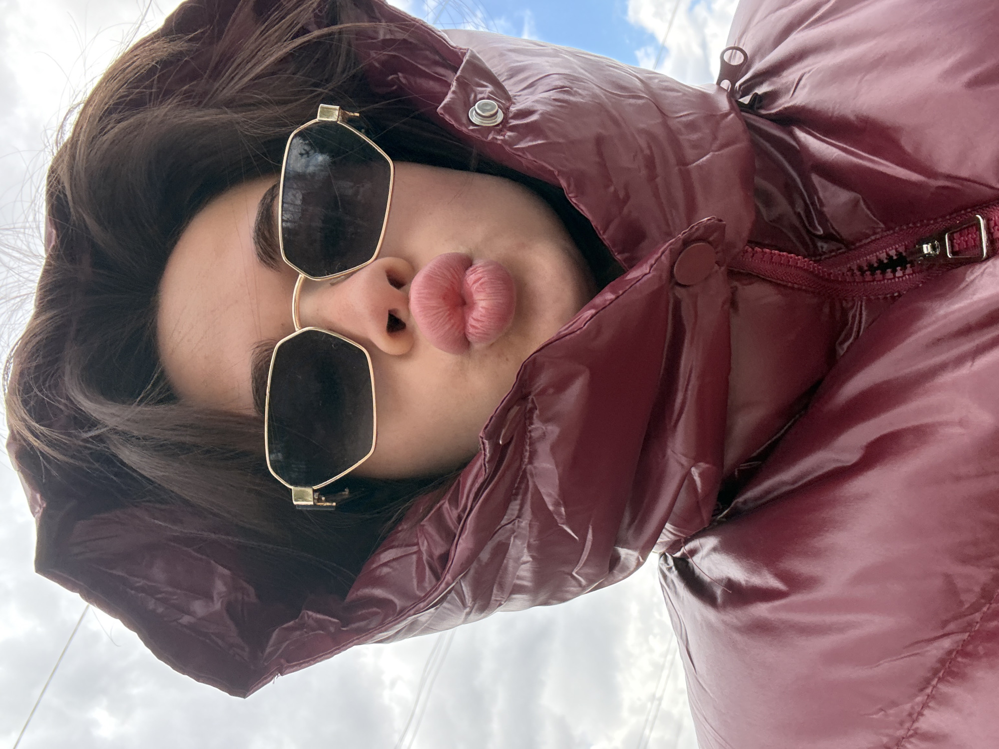 A low-angle selfie captures a person against a bright, cloudy sky, with the image rotated, suggesting the subject is looking down at the camera. The individual wears large, hexagonal sunglasses with dark lenses and gold-toned frames, obscuring their eyes. Their dark hair frames their face, which is largely encased by the hood of a shiny, puffy, dark red or maroon jacket.
A prominent feature is the person's pursed, pink lips, forming a "duck face" or pout, indicating a playful or stylized pose. The jacket, made of a glossy material, has visible details including a zipper with a metallic pull and snap buttons on the hood. The background shows an overcast sky with patches of blue and white clouds, signifying daytime.
No specific location cues are visible within the image itself, besides the sky. There is no visible text. The context places this image in Лобаново, Russia. The overall impression is a casual yet stylish outdoor portrait.