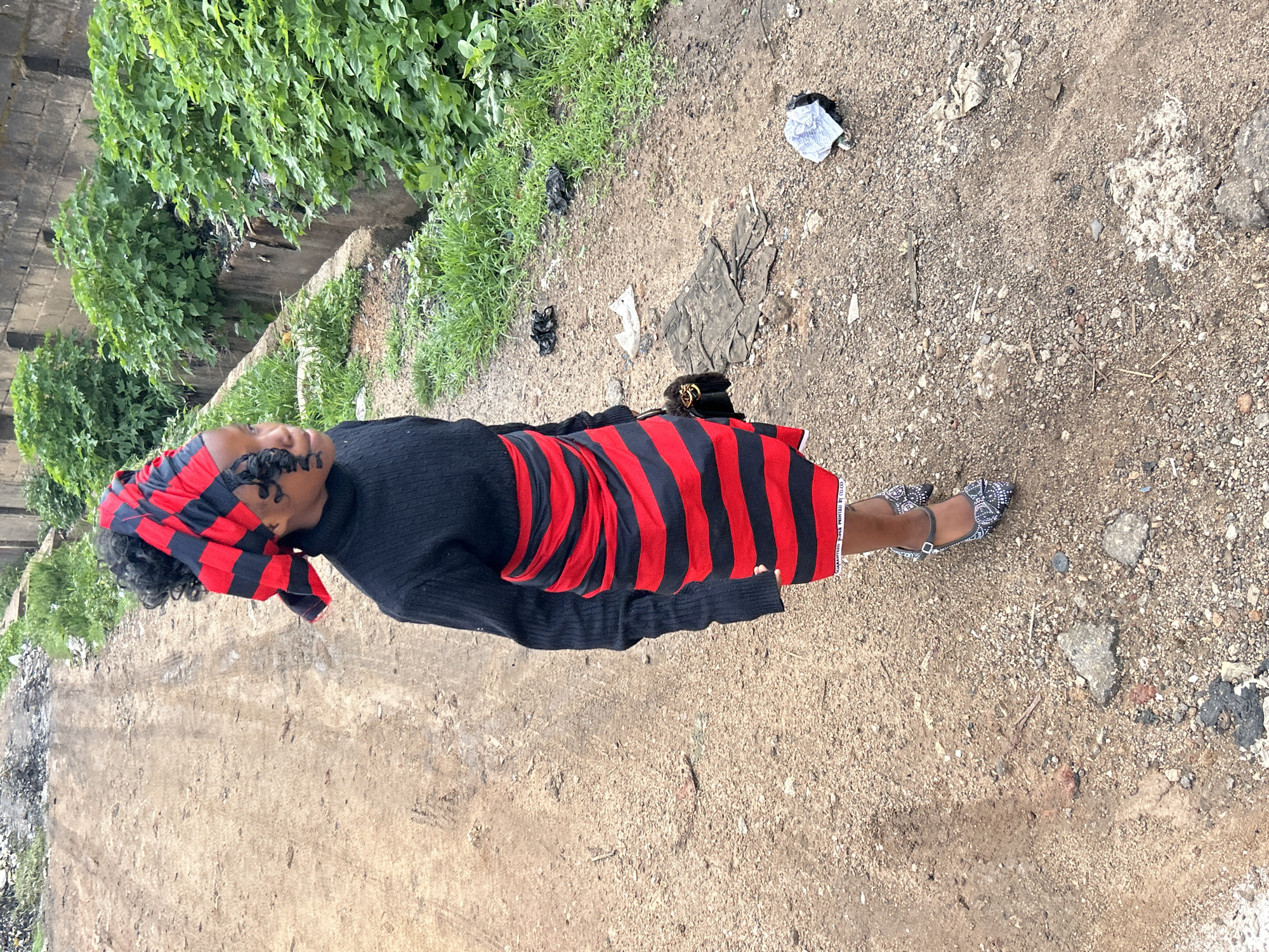 The image captures a young female standing on an unpaved dirt and gravel road in an urban environment, likely in Jos, Nigeria. The subject, centrally positioned and viewed from a slightly elevated angle, wears a black long-sleeved turtleneck top, a horizontally striped red and black wrap-around skirt, and a matching red and black striped headwrap. Her dark, curly hair is partially visible beneath the headwrap. She wears pointed-toe high-heeled shoes, which are grey or silver and adorned with decorative elements, secured by an ankle strap. A small black handbag with a gold-colored chain detail is visible on her left side. Her head is turned towards the right side of the frame.
The ground consists of coarse reddish-brown dirt and small stones, exhibiting areas of unevenness and scattered debris, including dark plastic fragments, pieces of paper, and small rock formations. To the right of the subject, a concrete drainage ditch runs parallel to the road, heavily overgrown with dense, vibrant green vegetation. Beyond the vegetation, a weathered concrete wall with rectangular indentations is discernible. The road extends into the background on the left, appearing similarly unpaved and showing signs of wear and scattered detritus. The lighting is soft and diffused, with no harsh shadows visible.