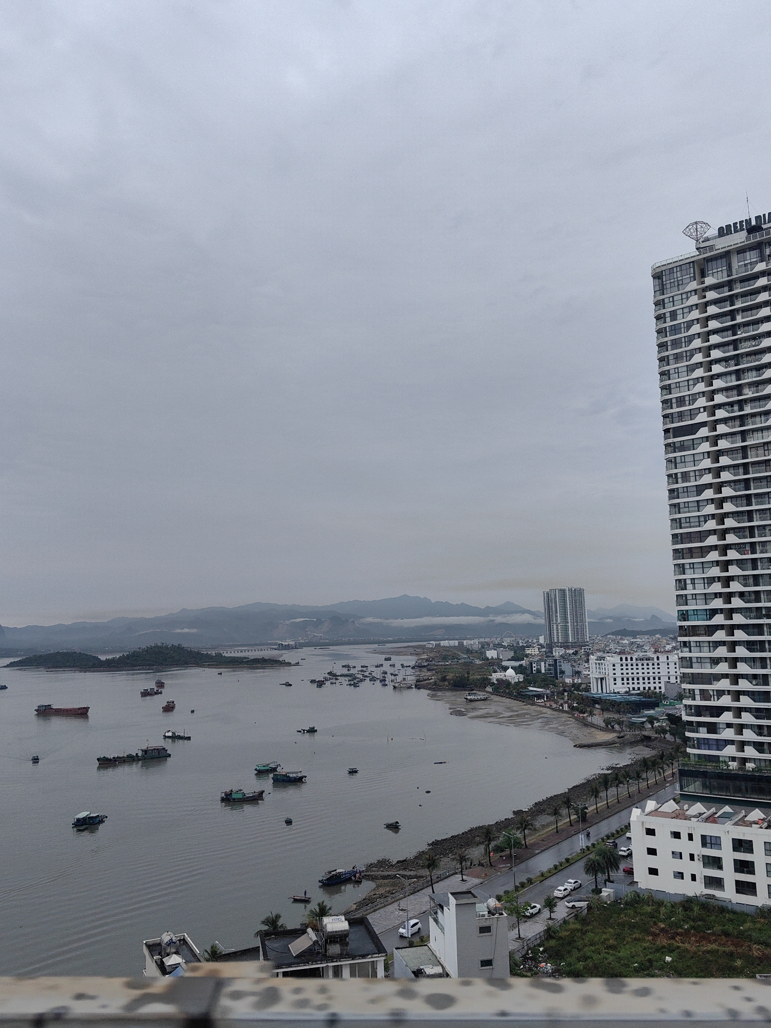 The image shows an elevated view of Hong Gai Ward, Vietnam on an overcast day. In the foreground is a section of railing from where the photograph was taken. To the right, a tall, modern high-rise building dominates the view. Further in the distance, along the coastline, are numerous smaller buildings. A coastal road runs along the edge of the bay, lined with palm trees and some vehicles. The bay itself is filled with boats of varying sizes. In the background, mountains are visible, obscured by the mist or fog. The sky is uniformly gray, casting a muted light across the entire scene.