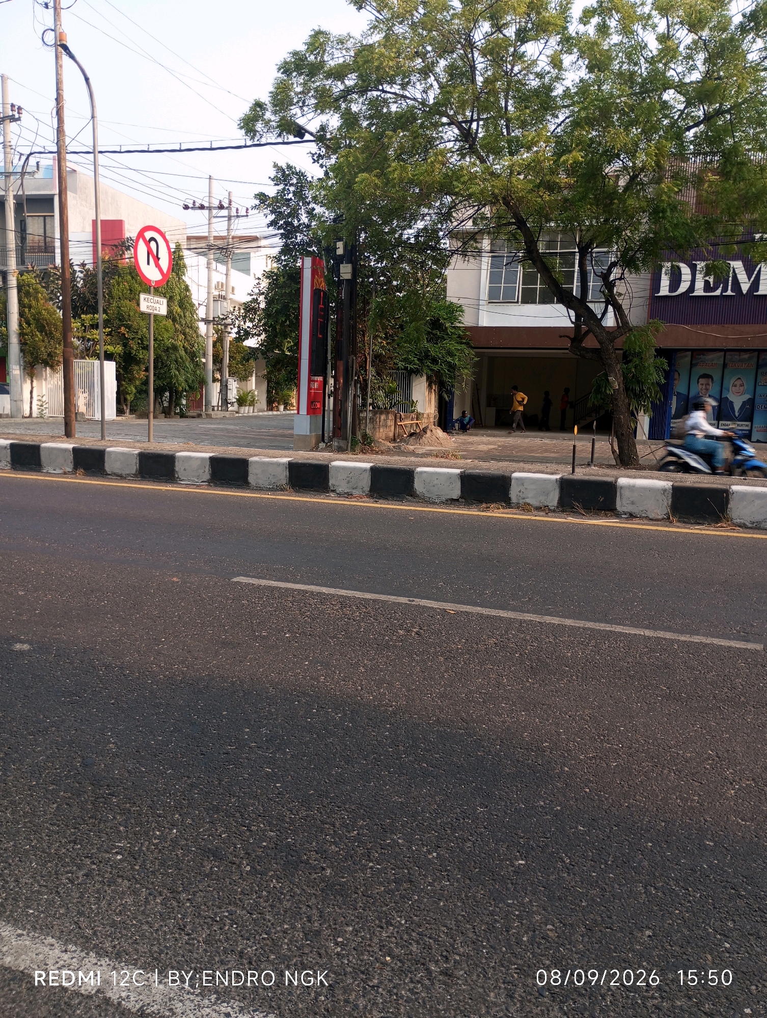 The image depicts a street scene in Ringinanom, Indonesia, on a clear day. A paved road with a yellow line and a white line runs horizontally across the foreground. The road is bordered by a curb with alternating black and white painted blocks. To the left of the road, a traffic sign prohibits U-turns, indicated by a red circle with a diagonal line over a U-turn arrow, with the text "KECUALI RODA 2" (except for two-wheeled vehicles) below it. Behind the sign, a tall utility pole with numerous wires is visible, along with trees and a building with white walls and windows.
On the right side of the road, a building with a purple sign that reads "DEM" is partially visible. In front of this building, there are several people, including a person on a blue motorcycle in motion, appearing to turn or move away from the camera. Trees with green foliage are present on the right side of the image, with one prominent tree in front of the building. The overall environment suggests a public street with commercial or residential buildings. The bottom of the image shows a timestamp: "08/09/2026 15:50".
