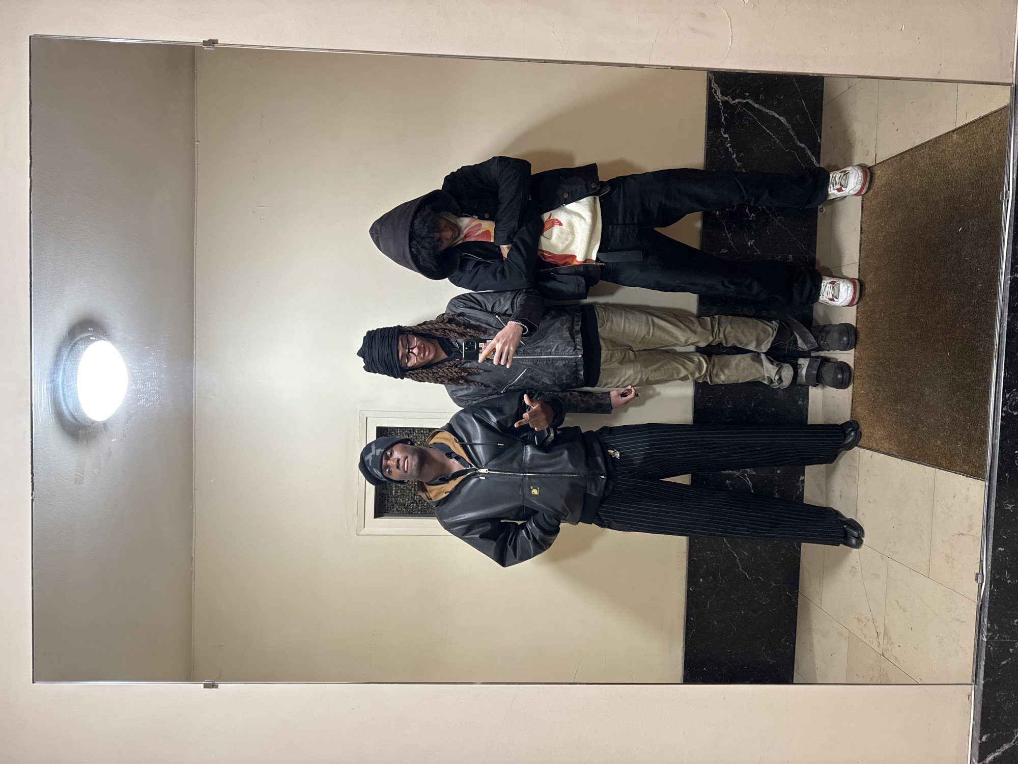The image depicts three male individuals reflected in a large mirror within an interior location in Paris, France. The scene appears to be a hallway or lobby, featuring light beige walls above a dark, polished marble-patterned dado rail. The floor consists of light-colored, possibly cream, square tiles, with a dark brown rectangular doormat positioned on top in the immediate foreground. A circular, flush-mounted light fixture is visible on the ceiling in the reflection, providing illumination.
From left to right in the reflection:
1. A male individual with dark skin wears a black beanie, a black leather-like bomber jacket over a brown collared shirt, and black pinstriped wide-leg trousers with dark shoes. His left hand is in his jacket pocket, and his right hand is raised with an open palm, fingers splayed. His mouth is slightly open, and he looks directly into the mirror/camera.
2. A second male individual, also with dark skin, wears a dark beanie with a subtle camouflage pattern, eyeglasses, a dark zippered jacket over a dark shirt, light brown trousers, and dark footwear with straps. He holds a dark-colored smartphone vertically with both hands, positioned to capture the mirror selfie. His gaze is directed towards the phone.
3. A third male individual, with dark curly hair visible, wears a black hooded jacket with the hood up, a cream-colored sweater with a multicolored abstract or graphic design underneath, black trousers, and white sneakers with red accents. His arms are crossed over his chest, and his head is tilted downwards, obscuring his direct gaze from the mirror/camera.
In the background reflection, positioned behind the leftmost individual's head, a rectangular inset panel containing a dark grate or vent is visible on the wall.