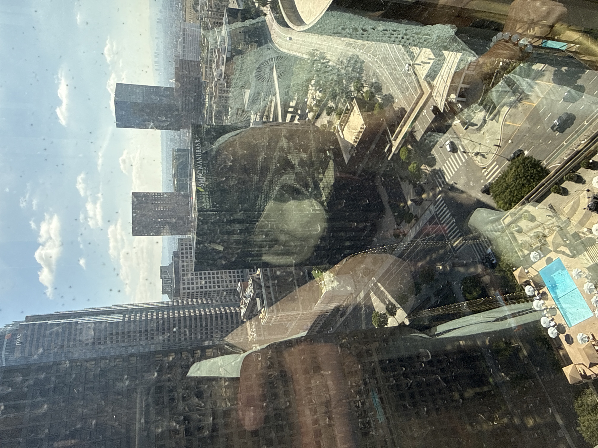 The image is a high-angle view of a cityscape, likely Los Angeles, California, United States of America, as seen through a dirty window. The window has numerous water spots and smudges, creating reflections and obscuring parts of the view. Several tall skyscrapers dominate the foreground and midground. One prominent building in the center has "SMBC MANUBANK" visible on its facade. To the left, a very tall, dark glass skyscraper with "pwc" logo is visible. In the distance, the city stretches out to the horizon under a bright, partially cloudy sky. Below the buildings, streets with cars and pedestrians are visible. A swimming pool with lounge chairs and umbrellas is situated on a lower rooftop. A hand with a bracelet is reflected in the window on the right side of the image. The overall impression is an urban environment on a sunny day.