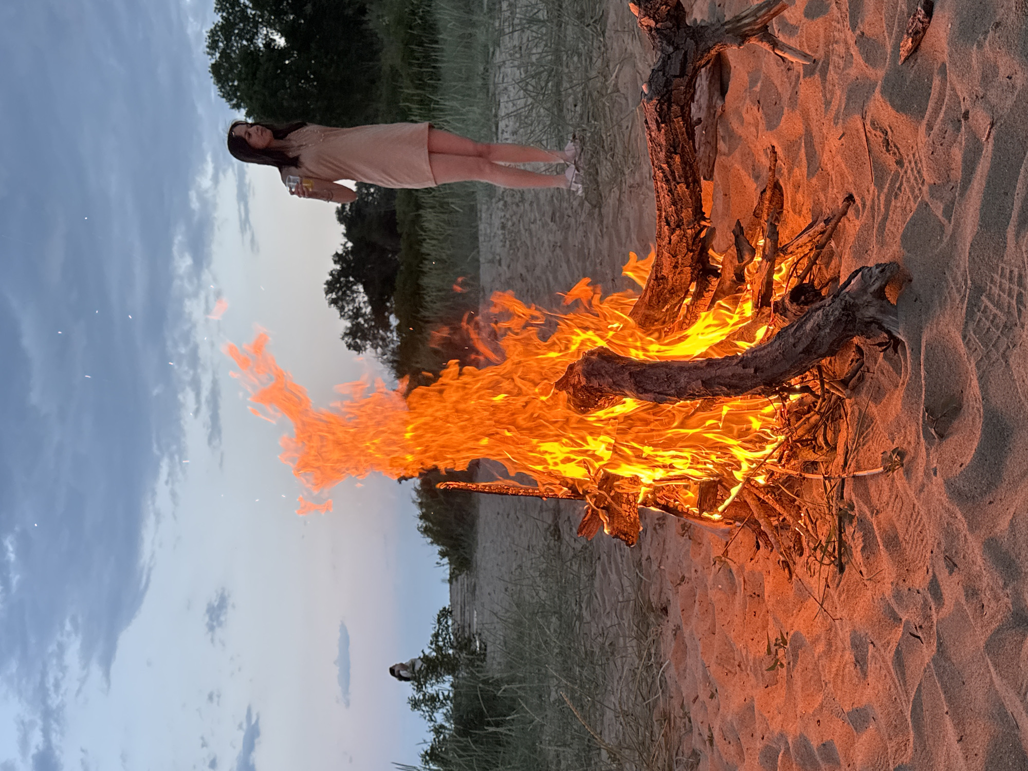 The image depicts an evening scene, likely at the beach in Novo Oriahovo, Bulgaria. A bonfire is prominently burning in the foreground on sand. To the left of the fire, a woman is standing with a drink in her hand. She wears a light-colored dress. Behind the woman and fire, there's some vegetation, possibly dune grass and trees, with the sky visible in the background. The sky has a gradient of colors, indicating either dusk or dawn. There is also a person visible behind the fire.