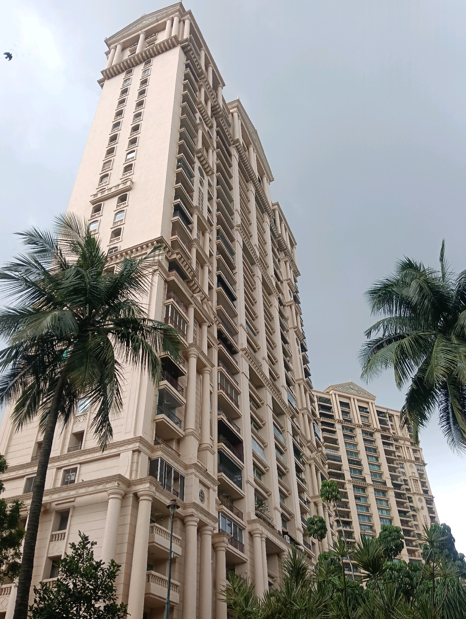 This media file depicts a low-angle view of a tall, cream-colored residential building with classical architectural elements, such as columns and ornate cornices. The building is modern, featuring numerous balconies and windows that suggest many apartments. In the foreground and to the right are lush green palm trees, characteristic of a tropical or subtropical climate. The sky is overcast and grey, indicating a cloudy or possibly rainy day. The overall atmosphere is serene and suggests an urban residential setting, possibly in a well-to-do area. There is a small dark bird flying in the sky to the left of the building. The presence of palm trees and the style of the building might suggest a location like Mumbai, India, known for its high-rise buildings and tropical climate. No people are visible in the media file. No text is visible.