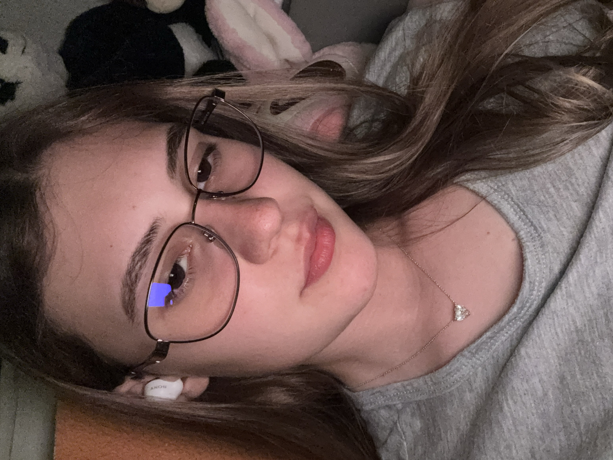 A close-up, rotated portrait shows a young person, likely female, lying down indoors in Panagyurishte, Bulgaria. The subject has fair skin and long, brown hair with lighter highlights, which frames her face. She wears thin, dark-framed eyeglasses; a clear blue rectangular reflection, possibly from a screen, is visible in the left lens. A white wireless earbud, bearing the brand "SONY," is tucked into her left ear.
She is dressed in a casual grey ribbed top, and a delicate necklace with a small, sparkling heart-shaped pendant rests on her collarbone. Her expression is neutral and calm, as she looks towards the camera. The background is softly blurred, revealing plush toys, including a pink bunny ear and a dark, fluffy object, suggesting a comfortable, personal space like a bedroom or living area. The lighting is soft and appears to be indoor ambient light, possibly from the evening. The image conveys a moment of relaxation.