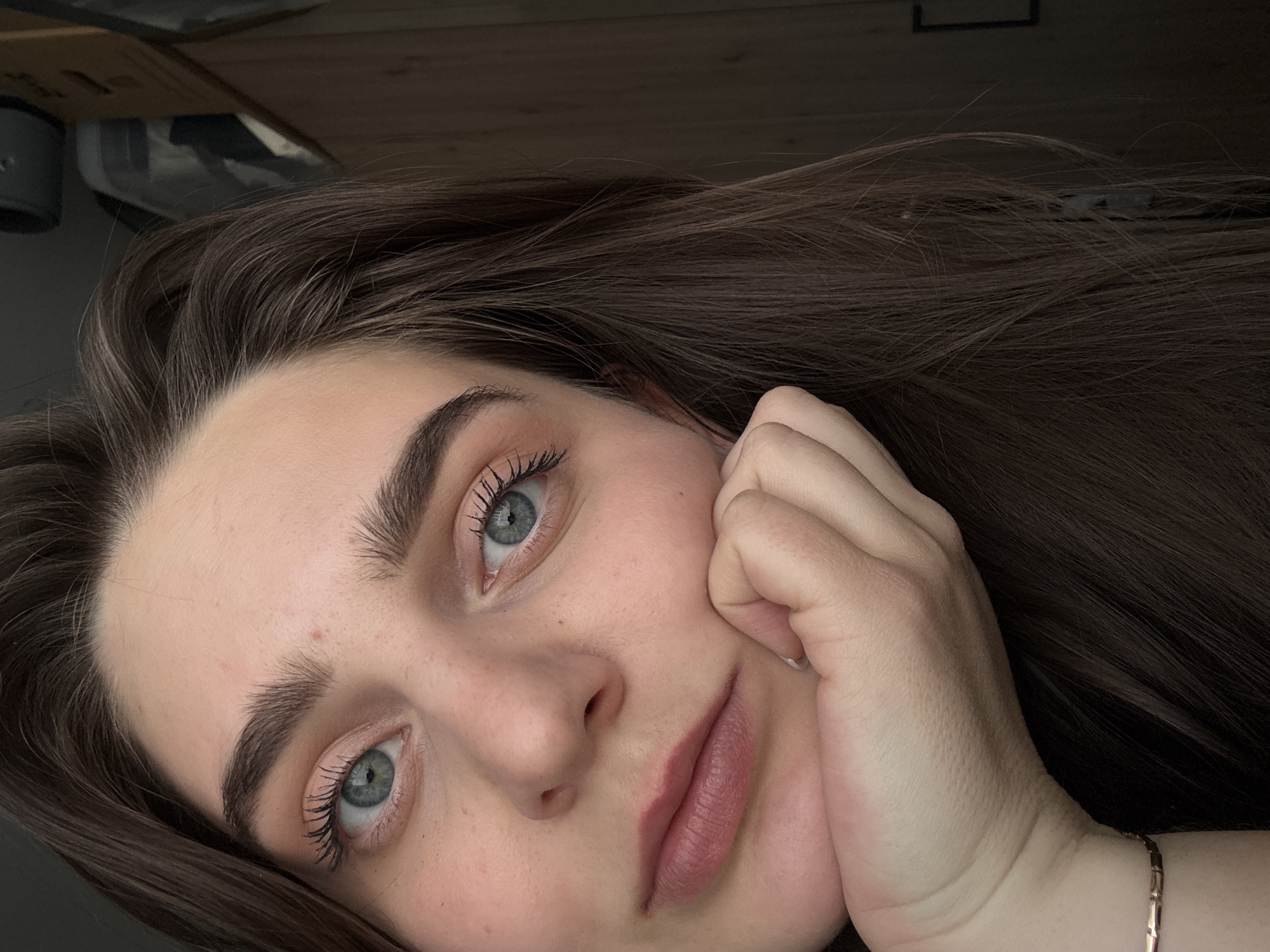 A close-up, tilted portrait captures a young woman with a calm and gentle expression, looking directly into the camera. Her head is angled towards the lower left of the frame. She has long, wavy dark brown hair that cascades around her. Her complexion is fair, and she features striking blue-green eyes framed by prominent dark eyebrows and long, dark eyelashes. A small, faint red blemish or freckle is visible on her forehead. Her lips are naturally colored with a subtle, rosy tint, forming a soft, almost imperceptible smile.
Her left hand, with neatly kept nails, supports her chin and cheek, partially covering the right side of her face. A slender, rose gold-colored bracelet with small rectangular links adorns her wrist. The background is dark and indistinct, suggesting an indoor setting with blurred, dark objects (possibly furniture or boxes) and a dark wall or ceiling. The lighting is soft and ambient, highlighting her facial features without harsh shadows. The image has a serene and intimate atmosphere. No visible text is present in the image.