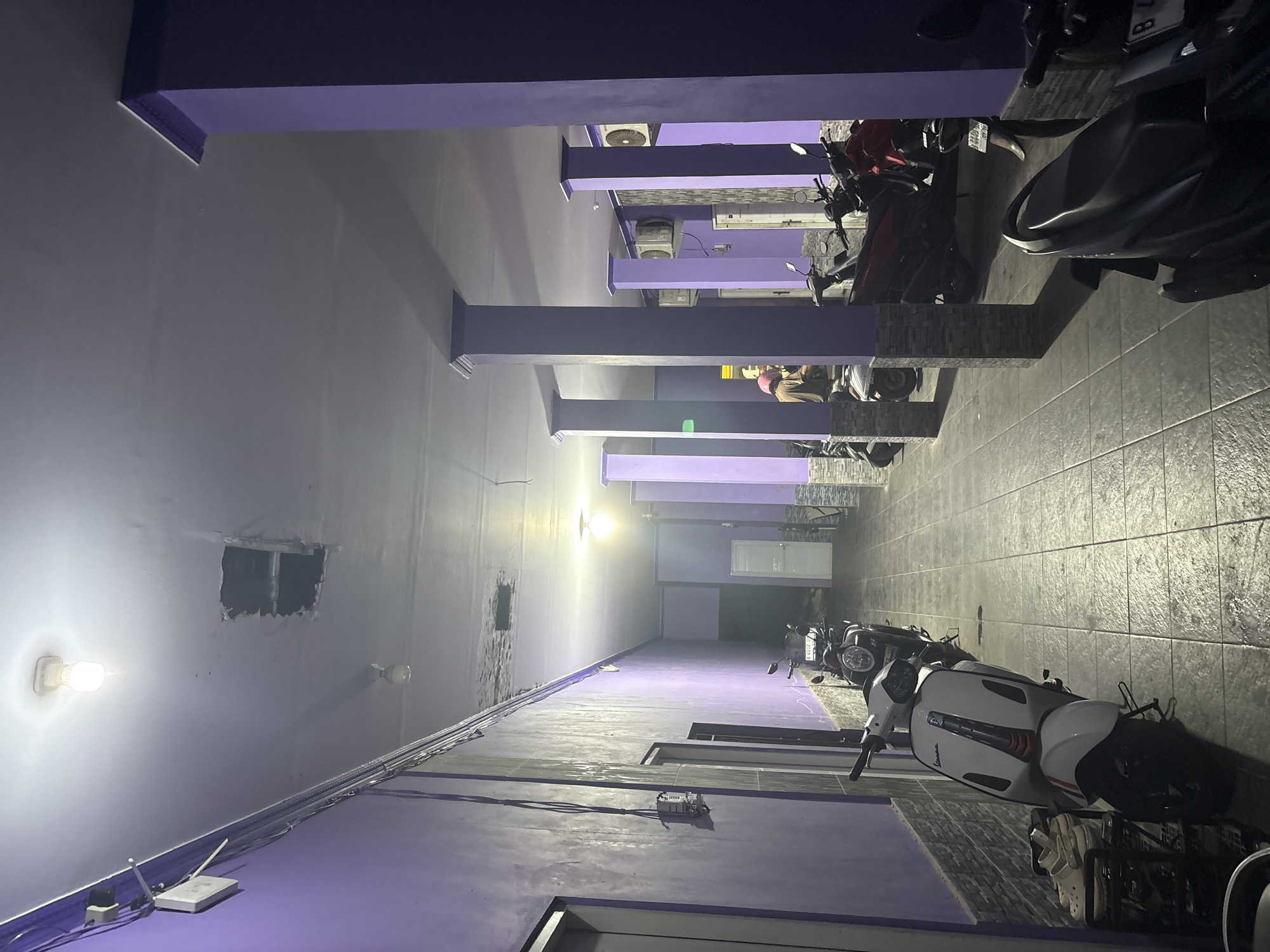 The image depicts a long, narrow hallway with purple-painted walls, illuminated by overhead lights. The floor is tiled with square grey tiles. Several motorcycles are parked along the right side of the hallway. In the foreground, a white Vespa scooter is parked. Further down the hallway, other motorcycles, including a red one, are visible. On the left side of the hallway, there are doors and electrical conduits running along the wall. The ceiling has some water damage and a hole. The scene appears to be an indoor common area or corridor, possibly in a residential or commercial building. No people are clearly visible or engaged in any specific action. The image does not contain enough information to identify the specific location in North Jakarta, Indonesia.