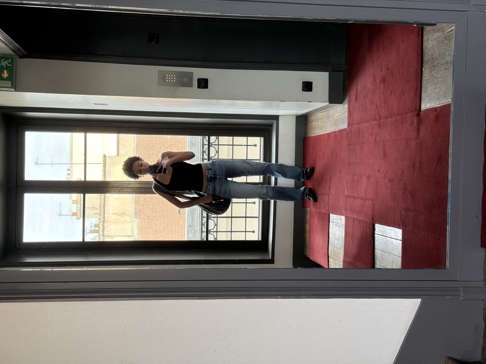 A young woman with short, curly hair stands in front of a window, taking a mirror selfie. She is wearing a black tank top, blue jeans, and black shoes. She has a dark bag slung over her shoulder. The window behind her looks out onto a Parisian street with buildings and rooftops visible. A red carpet is laid out on the floor in front of the mirror. The scene appears to be an indoor hallway or entryway.